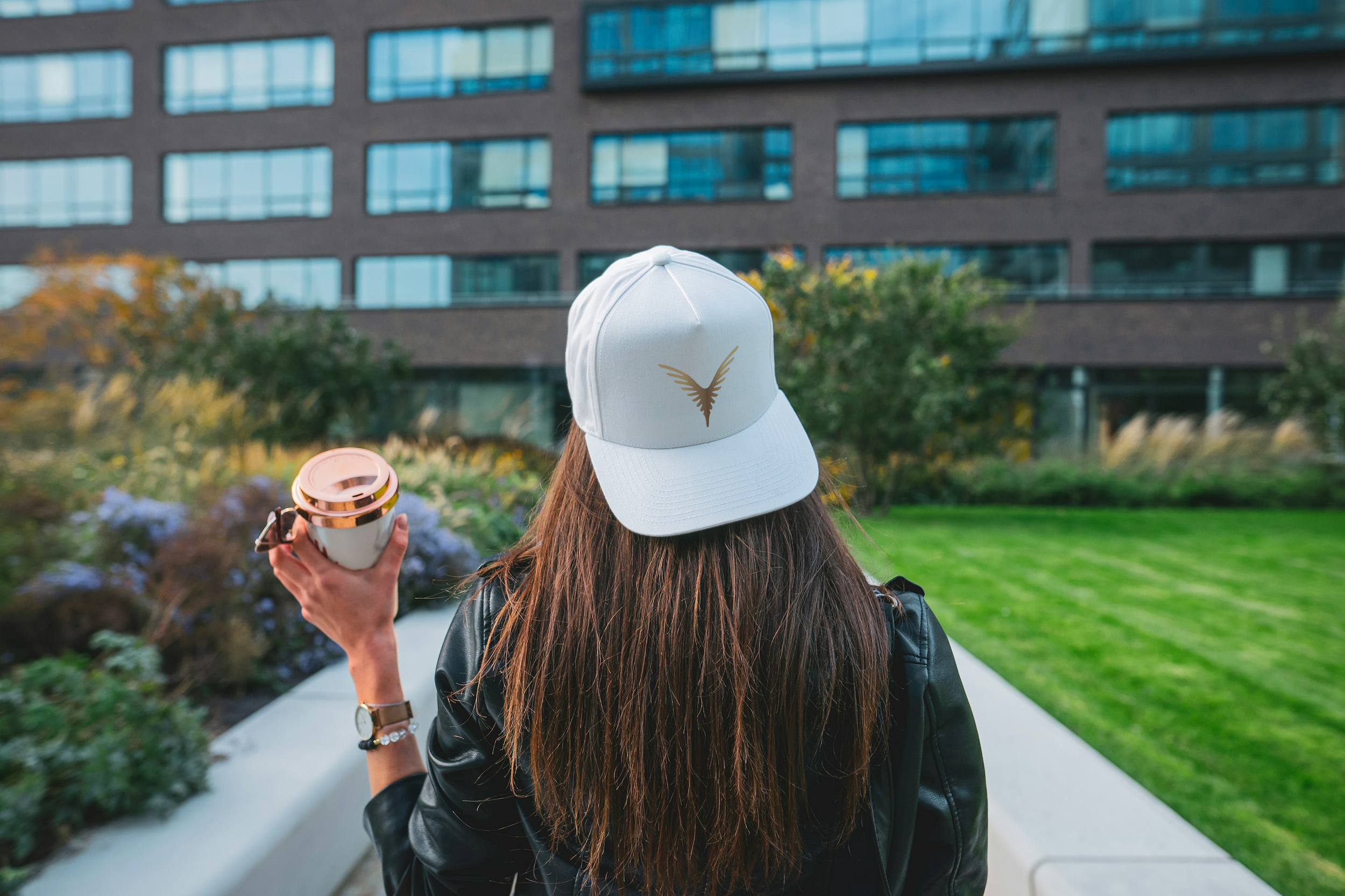 Femme de dos portant une casquette blanche avec un logo d'oiseau doré, tenant une tasse à café dans un jardin urbain avec un bâtiment moderne en arrière-plan.