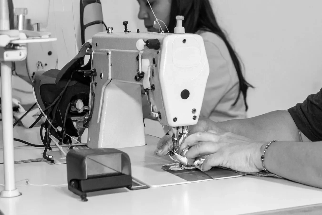 Close-up of hands guiding fabric through a sewing machine in a workspace, with a woman observing in the background.