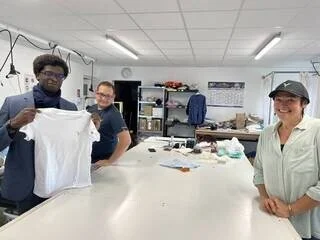 Three people in a workspace holding a white t-shirt and smiling, with shelves, tables, and office supplies in the background.