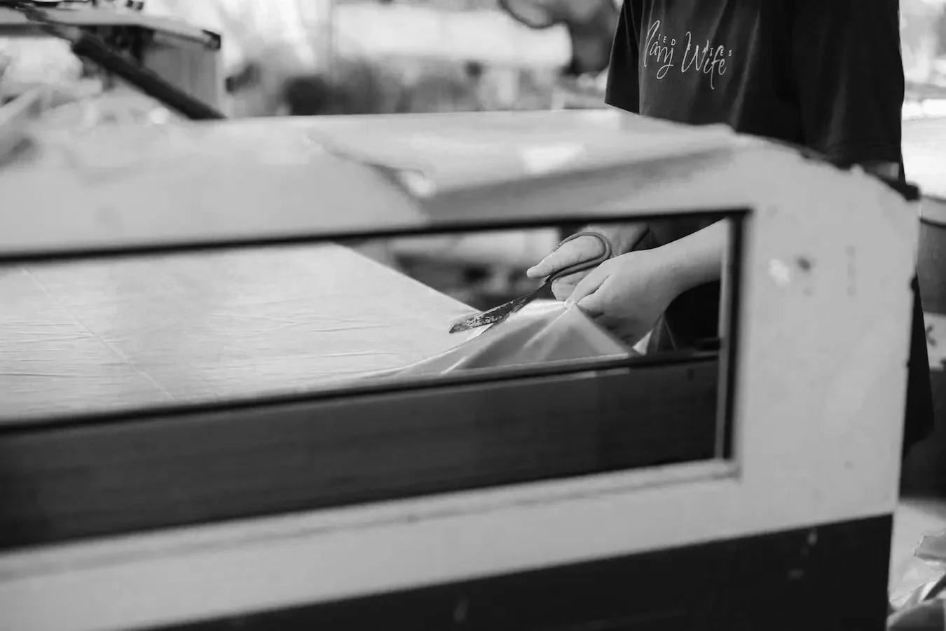 Hands cutting a sheet of paper or fabric with scissors inside a display case or oven, with a person wearing a dark shirt nearby.