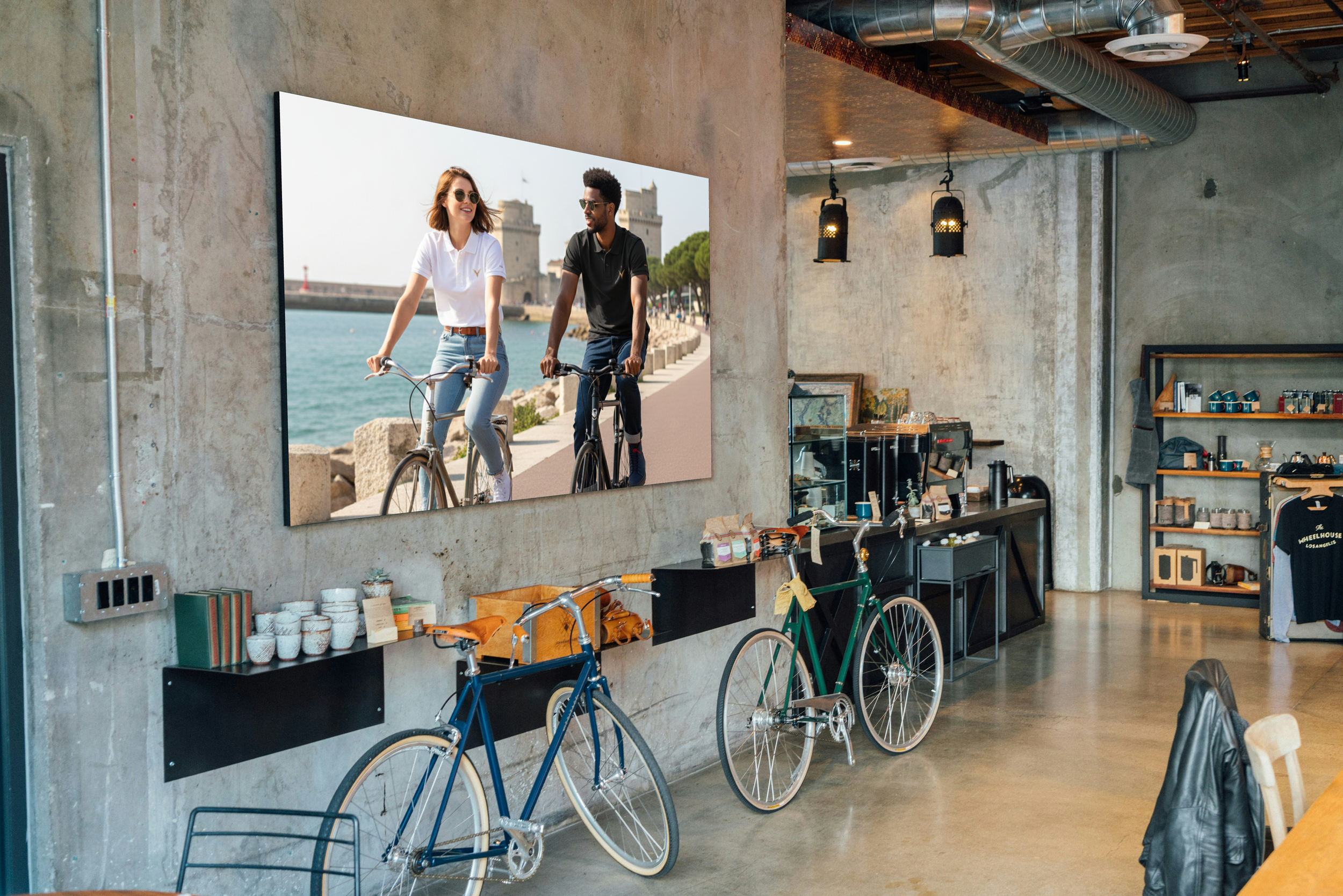Intérieur d'un café avec des vélos décoratifs, une grande affiche de deux personnes à vélo près de l'eau, étagères avec des boissons, vaisselle, et des objets décoratifs.