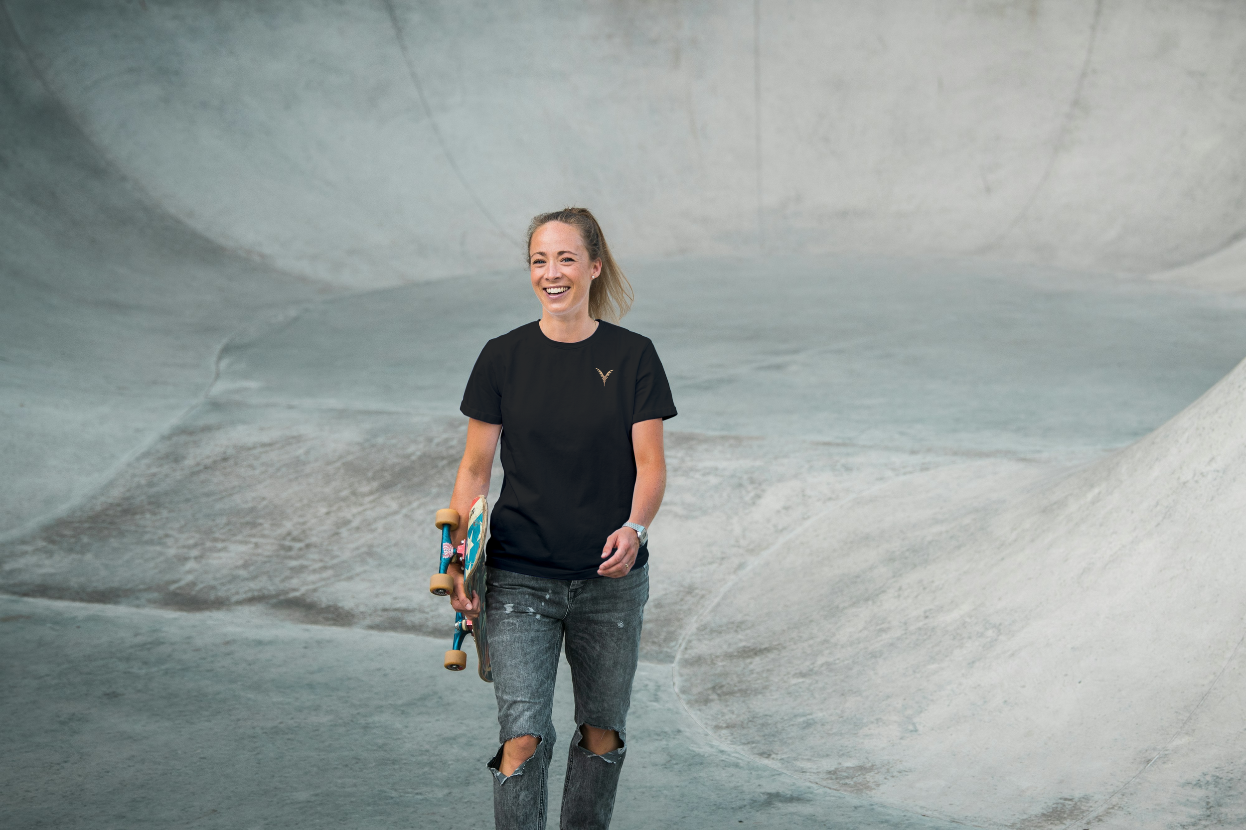 Femme souriante qui marche dans un skatepark en béton, tenant un skateboard dans sa main droite.