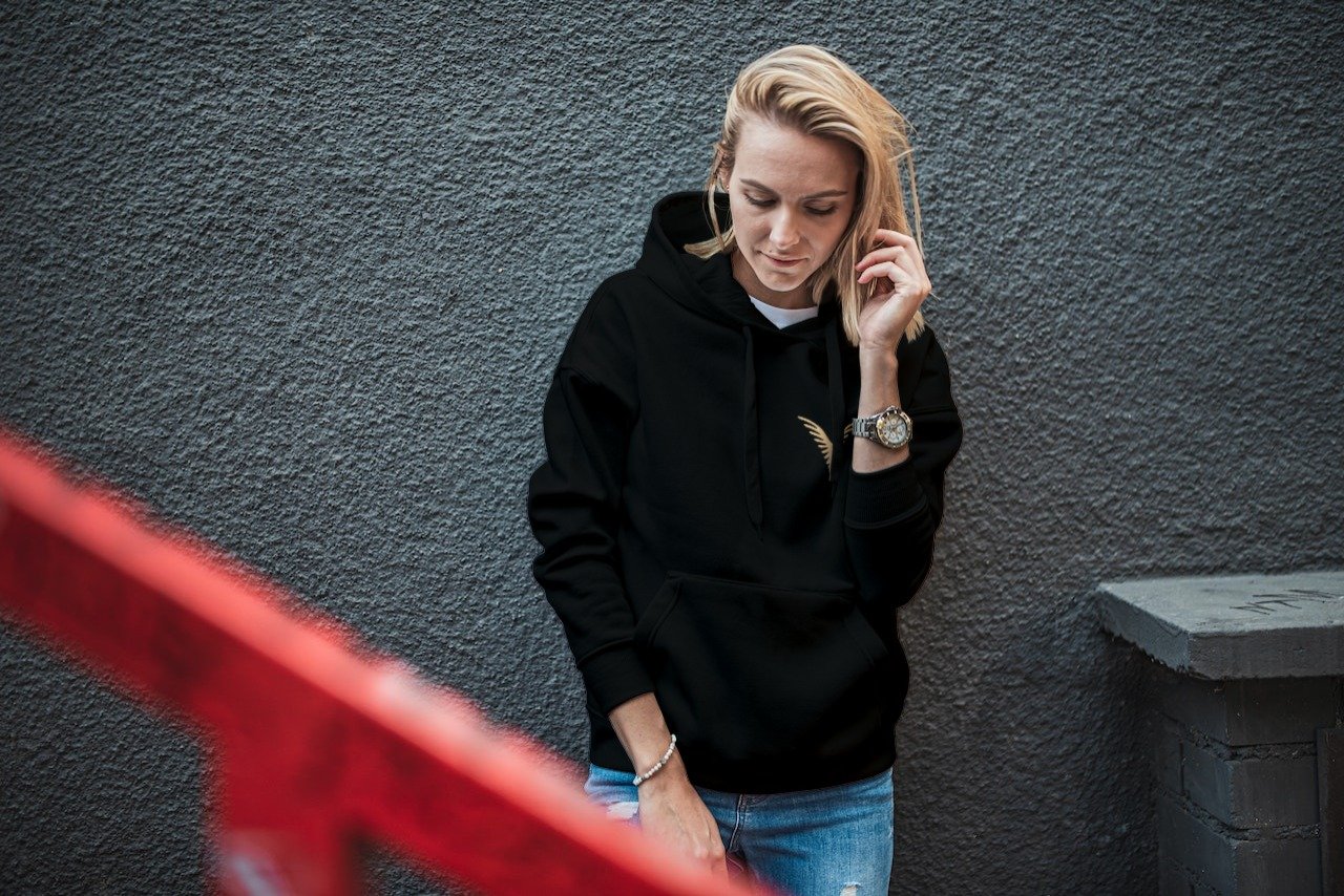 Une femme blonde portant un sweat à capuche noir, un jean bleu et une montre en bracelet en métal, se tient contre un mur gris, semblant réfléchir ou préoccupée, avec un bras dans la poche et l'autre touchant sa joue.