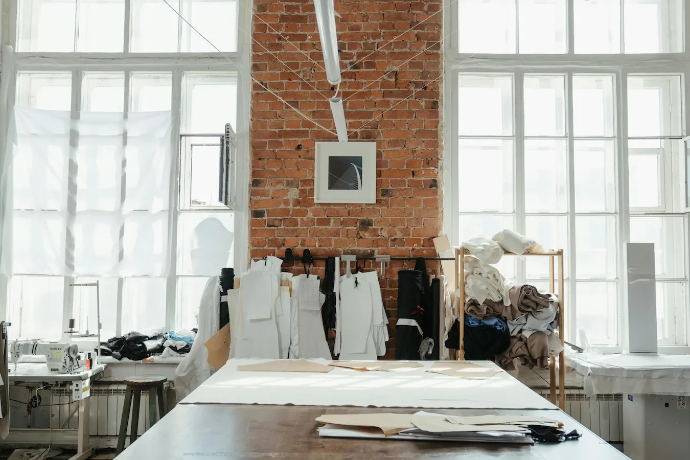 A well-lit sewing atelier with large windows, sewing machines, hanging white and black fabric pieces, shelves with folded fabric, and a brick wall with artwork.