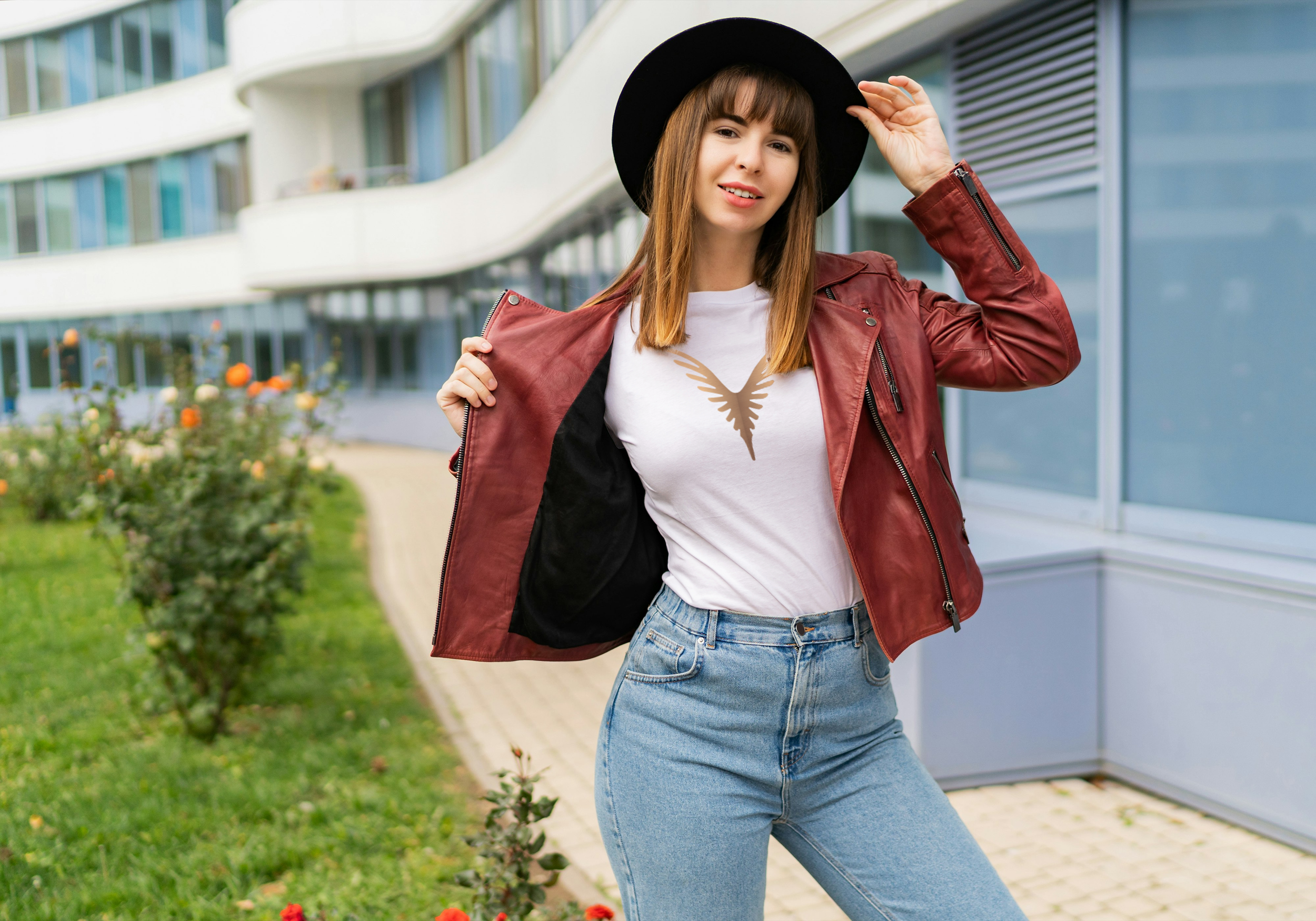 Jeune femme souriante portant un chapeau noir, une veste rouge en cuir, un t-shirt blanc à motif d'oiseau, et des jeans, debout devant un bâtiment moderne en verre.