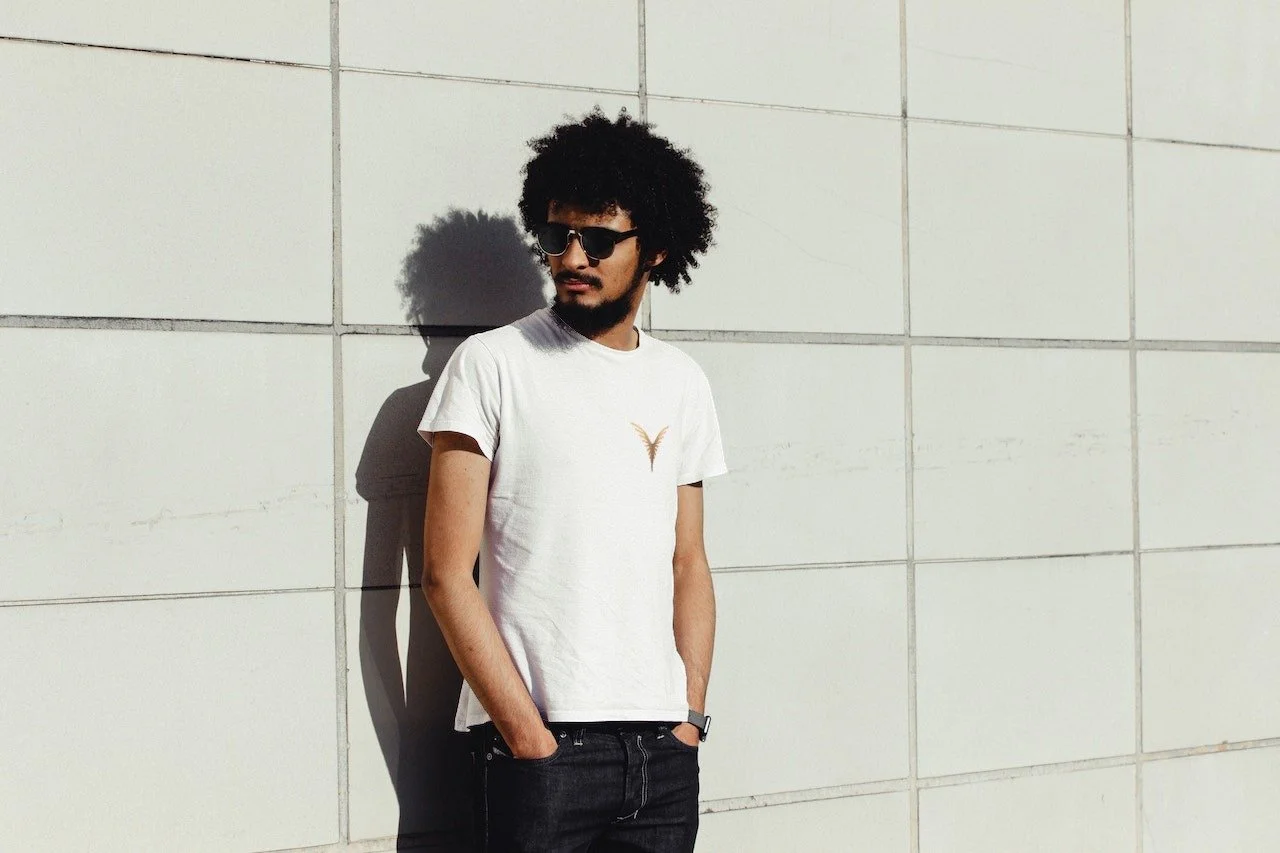 Un homme avec des cheveux afro portant des lunettes de soleil, une T-shirt blanc avec un petit logo en forme de V, et un pantalon noir, debout contre un mur blanc sous le soleil.