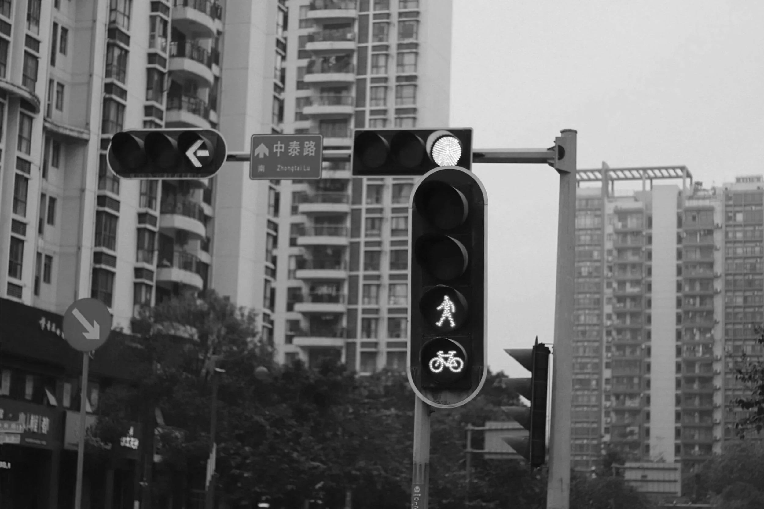 A black and white image of a traffic light showing a green pedestrian signal and a green bicycle signal, with tall apartment buildings in the background.