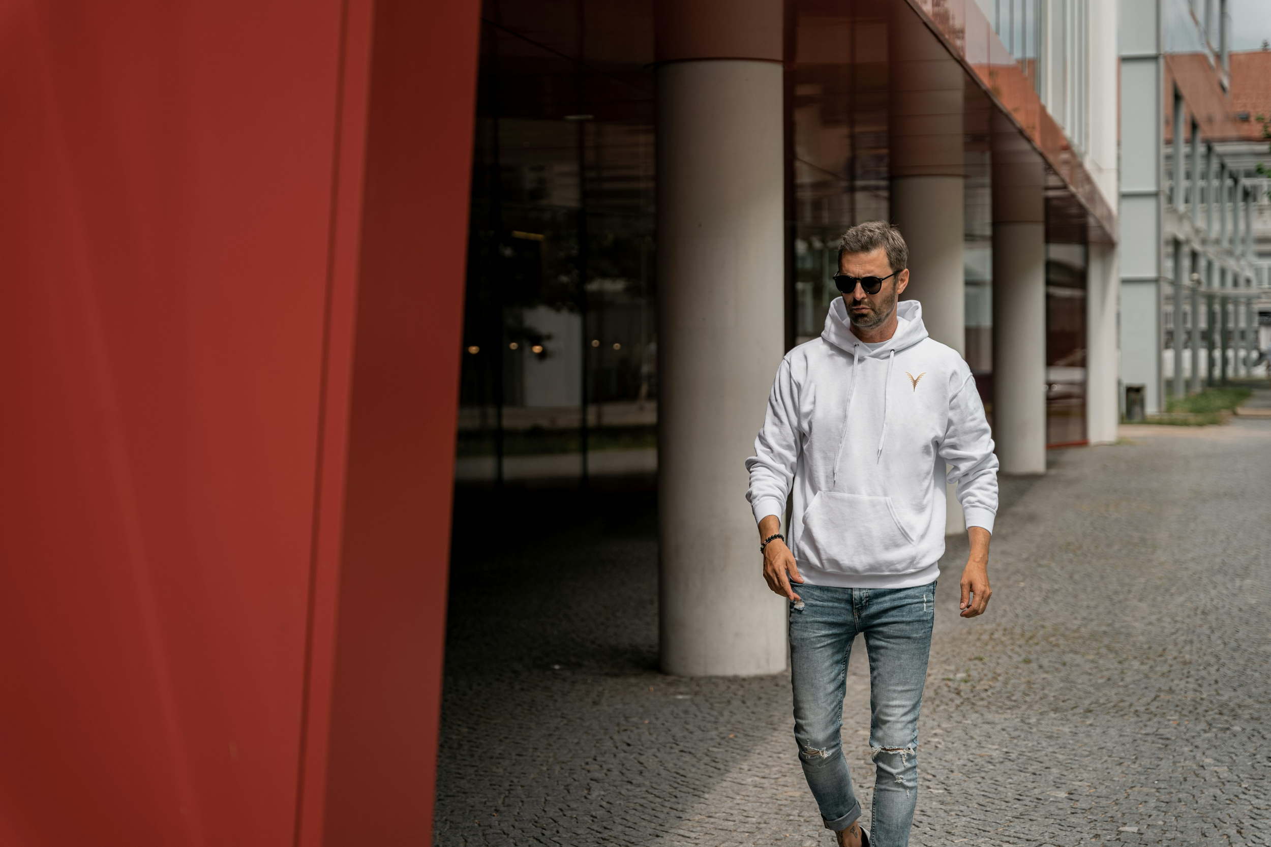 Un homme portant un sweat à capuche blanc et des lunettes de soleil marche à côté d'une structure moderne en ville.