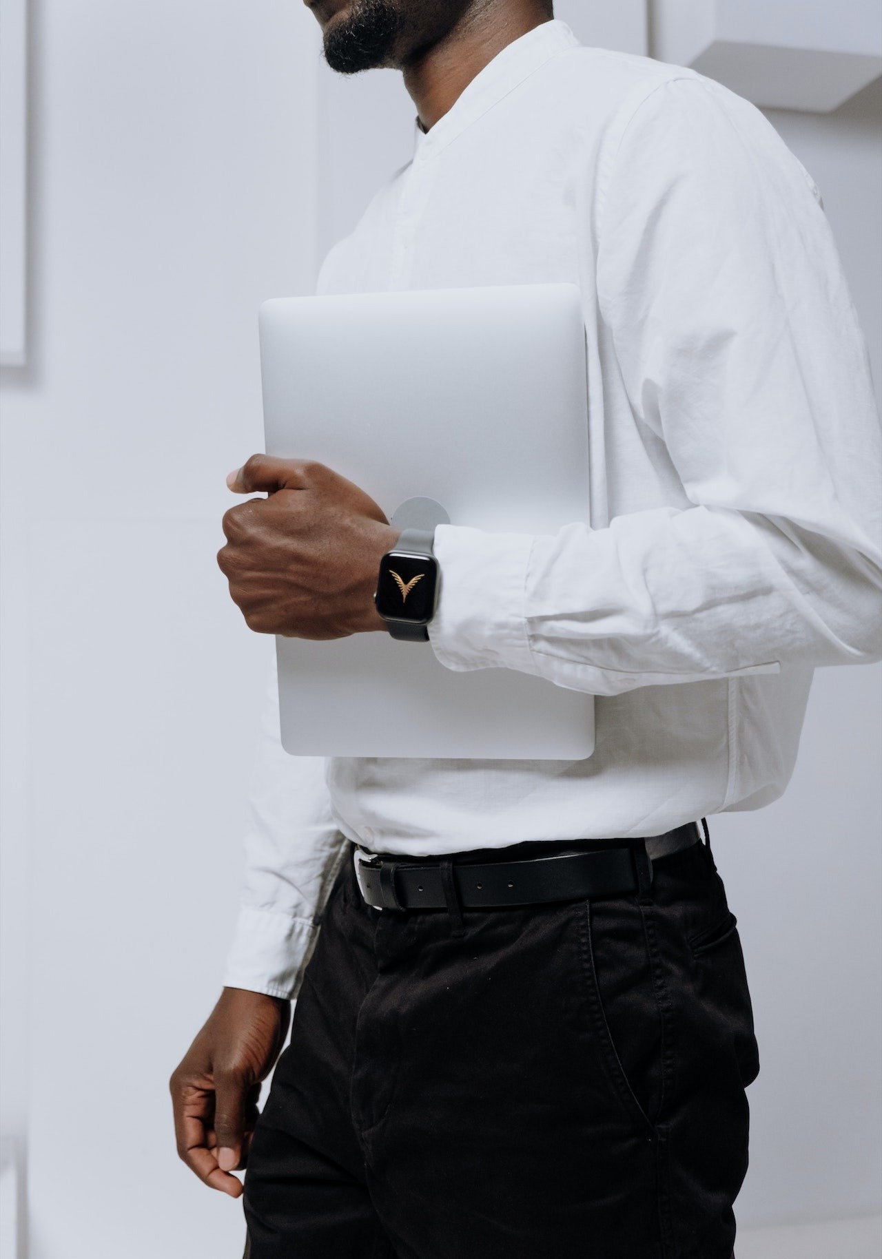A man wearing a black smartwatch displaying a V-shaped logo, holding a closed silver laptop, dressed in a white shirt and black pants in an office setting.