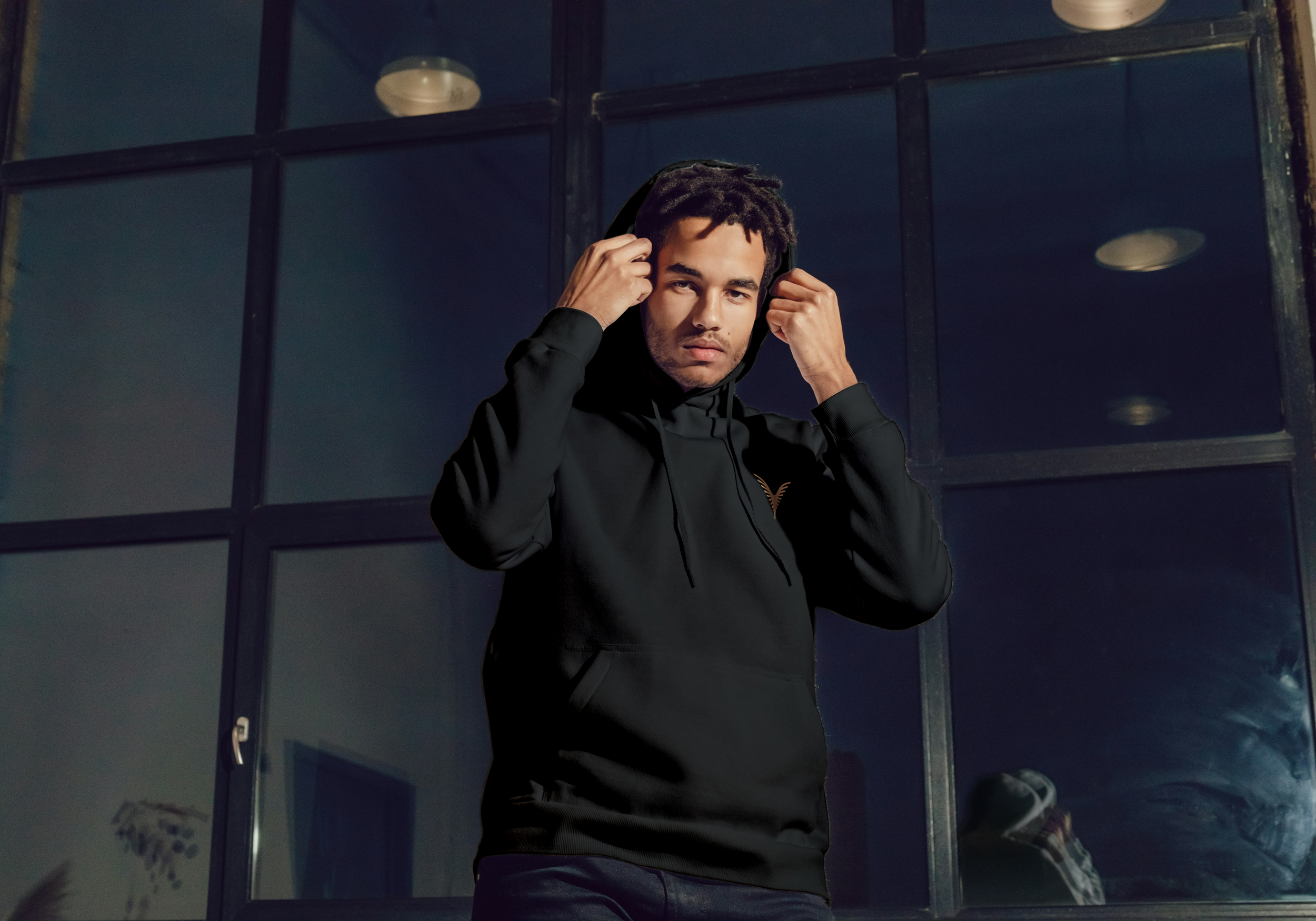 Un jeune homme porte un hoodie noir avec des écouteurs, devant une grande fenêtre en verre.