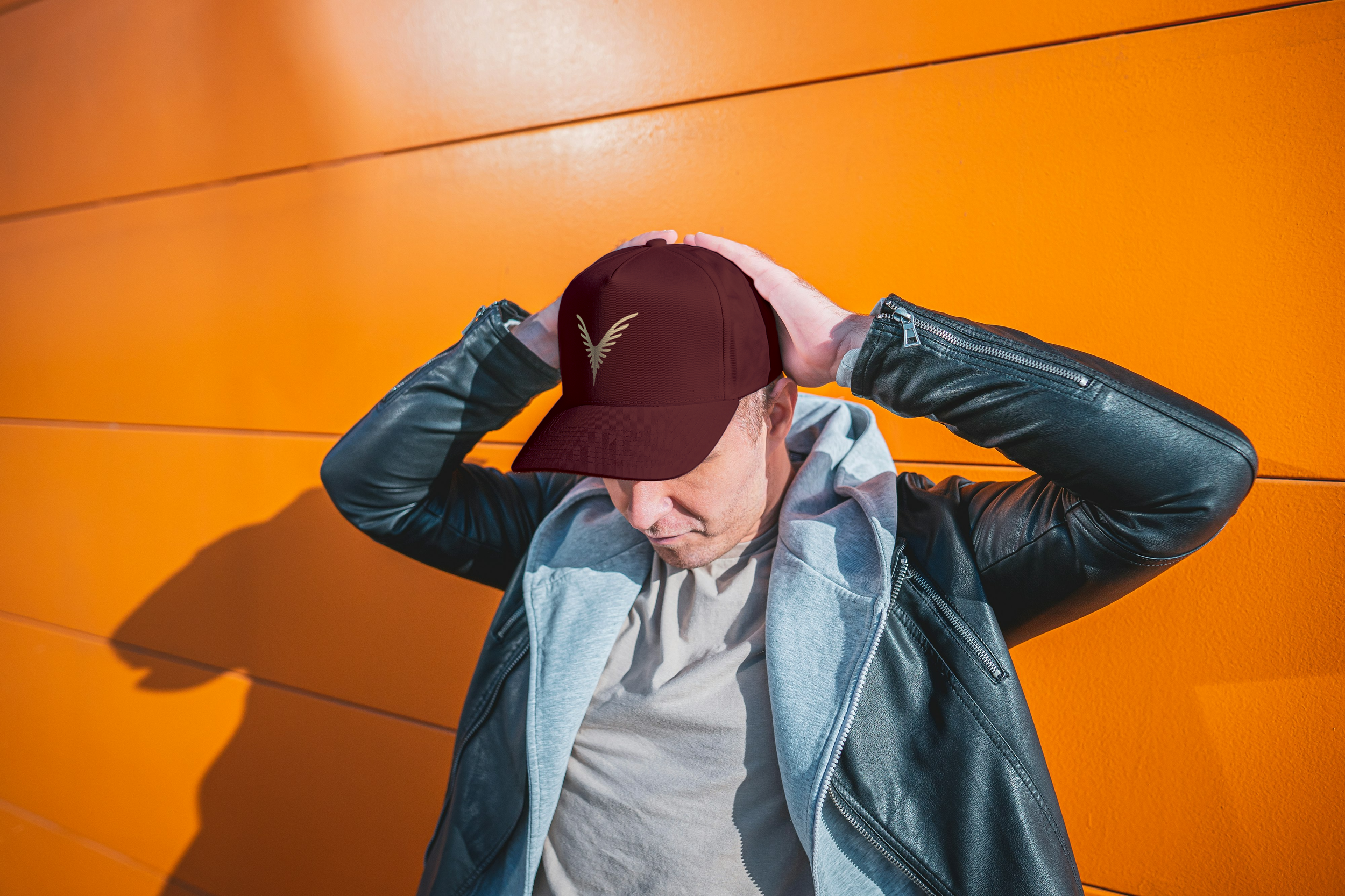Homme portant une casquette noire, une veste en cuir noire et un sweat gris, debout devant un mur orange, les mains sur la tête.