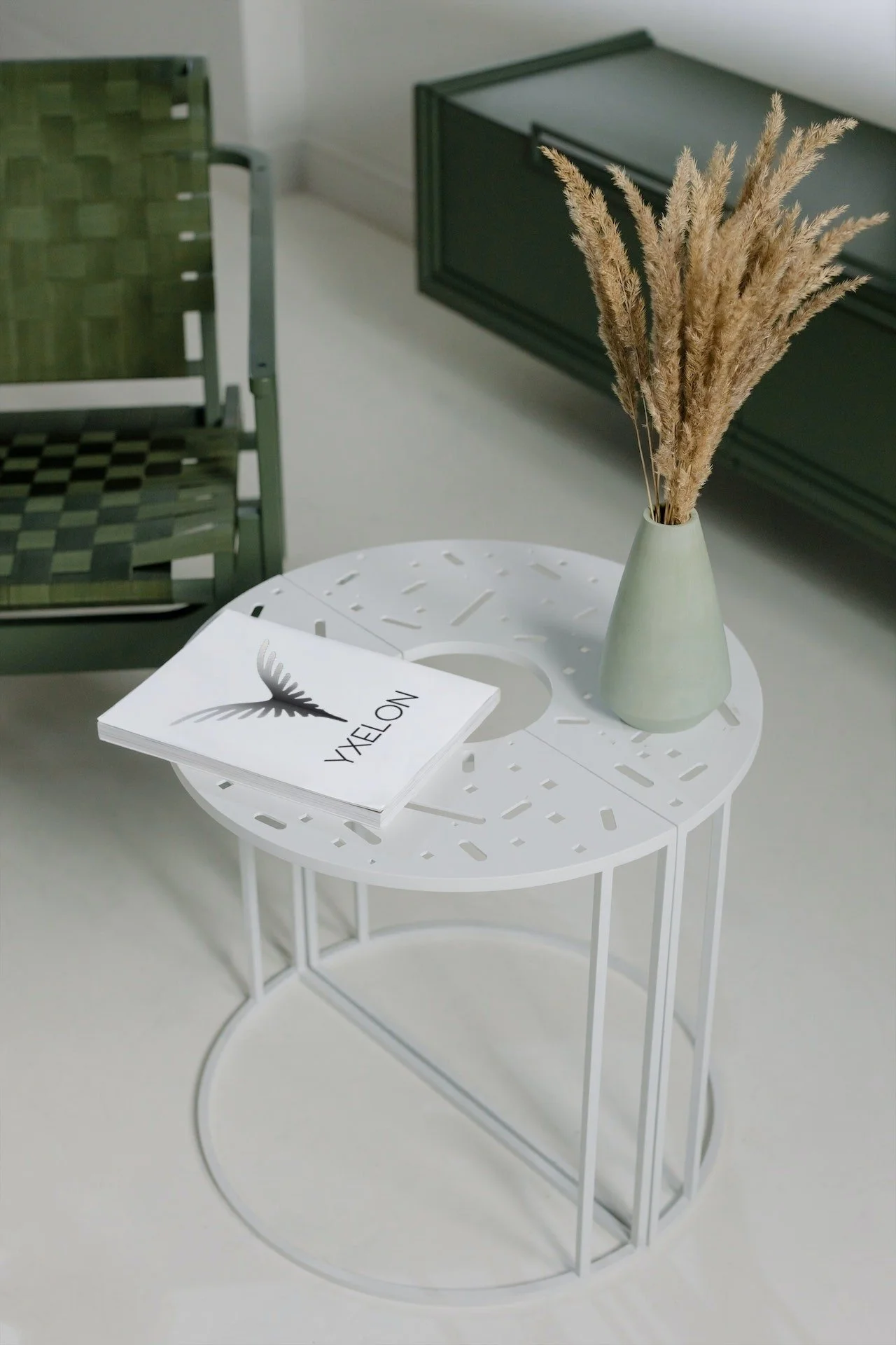 Une table basse ronde blanche avec un vase beige contenant des plantes séchées et un carnet blanc avec le logo YXELON gris posé dessus, dans un intérieur moderne minimaliste.