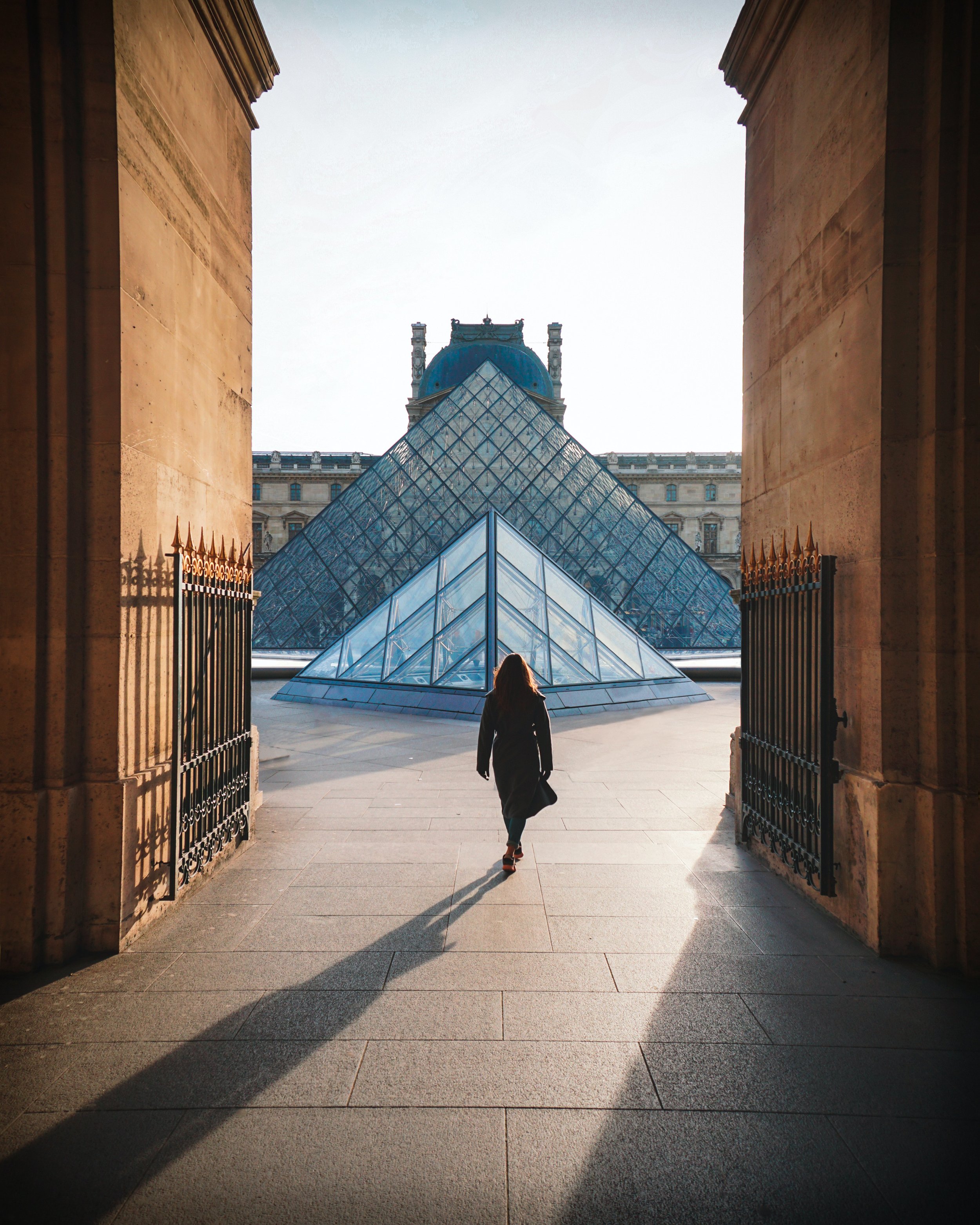 A woman walking towards the glass pyramid entrance of the Louvre Museum in Paris, France, as sunlight casts shadows on the ground.