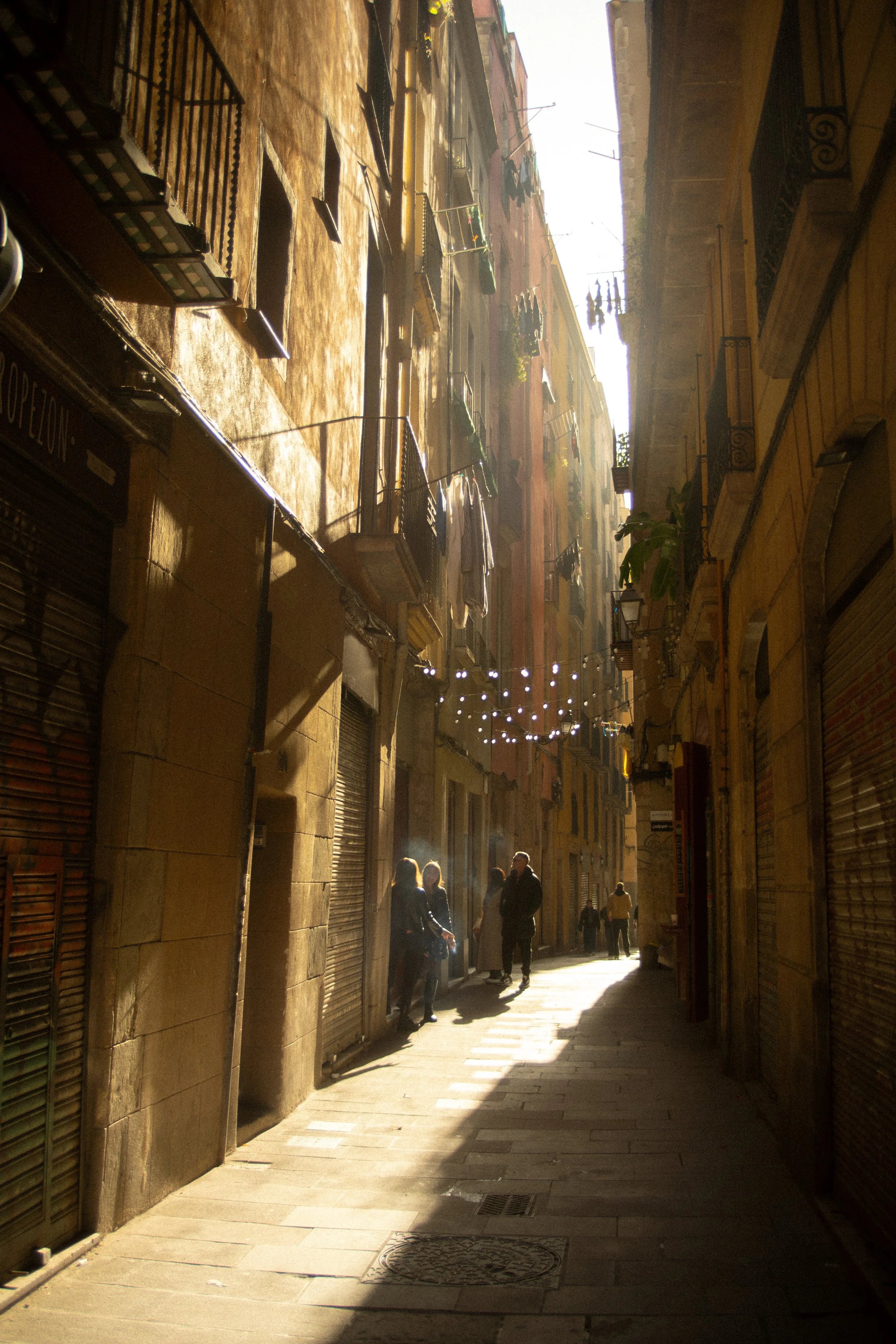 Sunlit narrow street with closed shop shutters, string lights, and a few people walking.