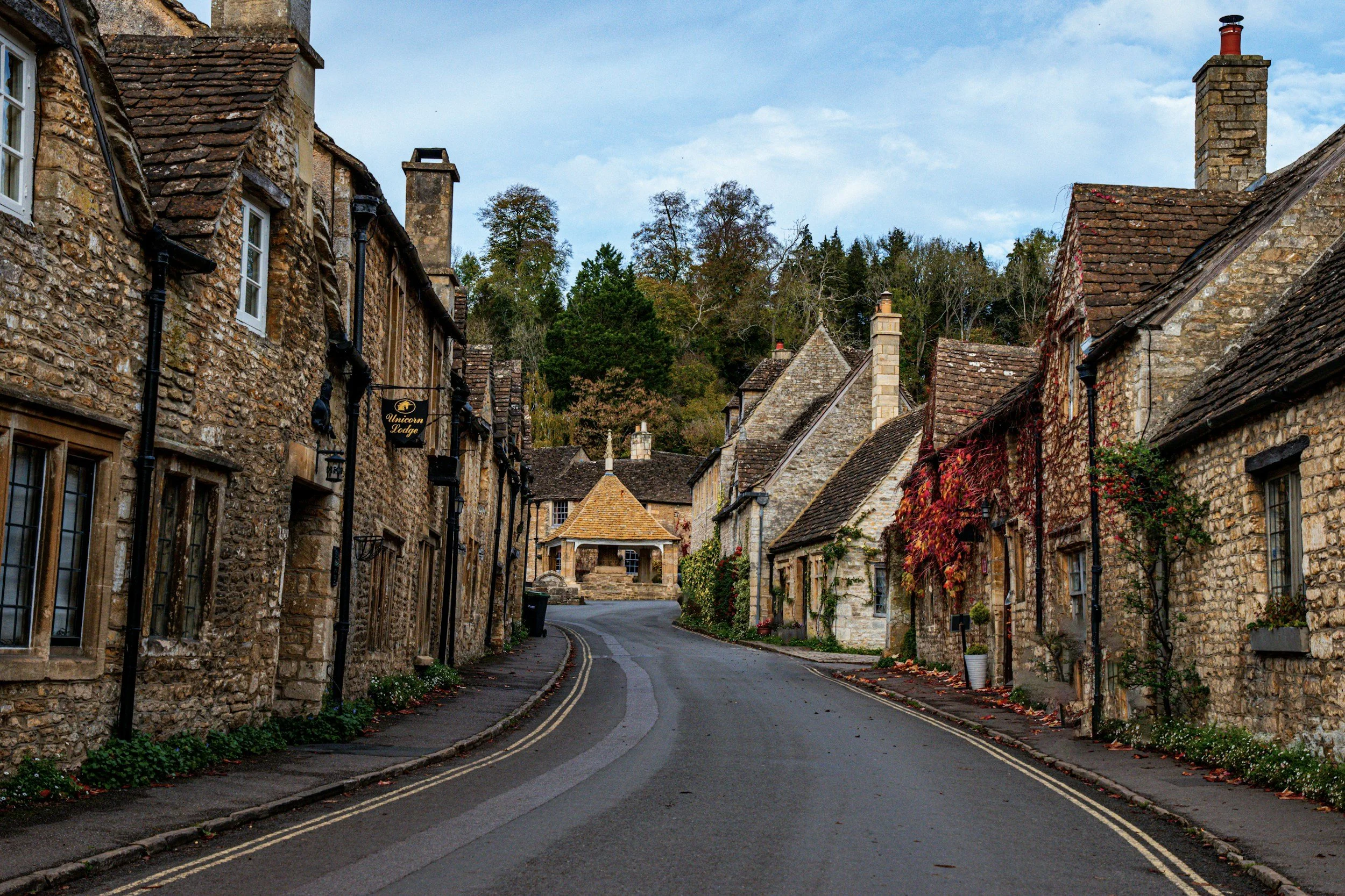 A narrow street in a small village with old stone houses on both sides, some adorned with climbing red and green plants, leading to a pavilion at the end, with trees and a cloudy sky in the background.