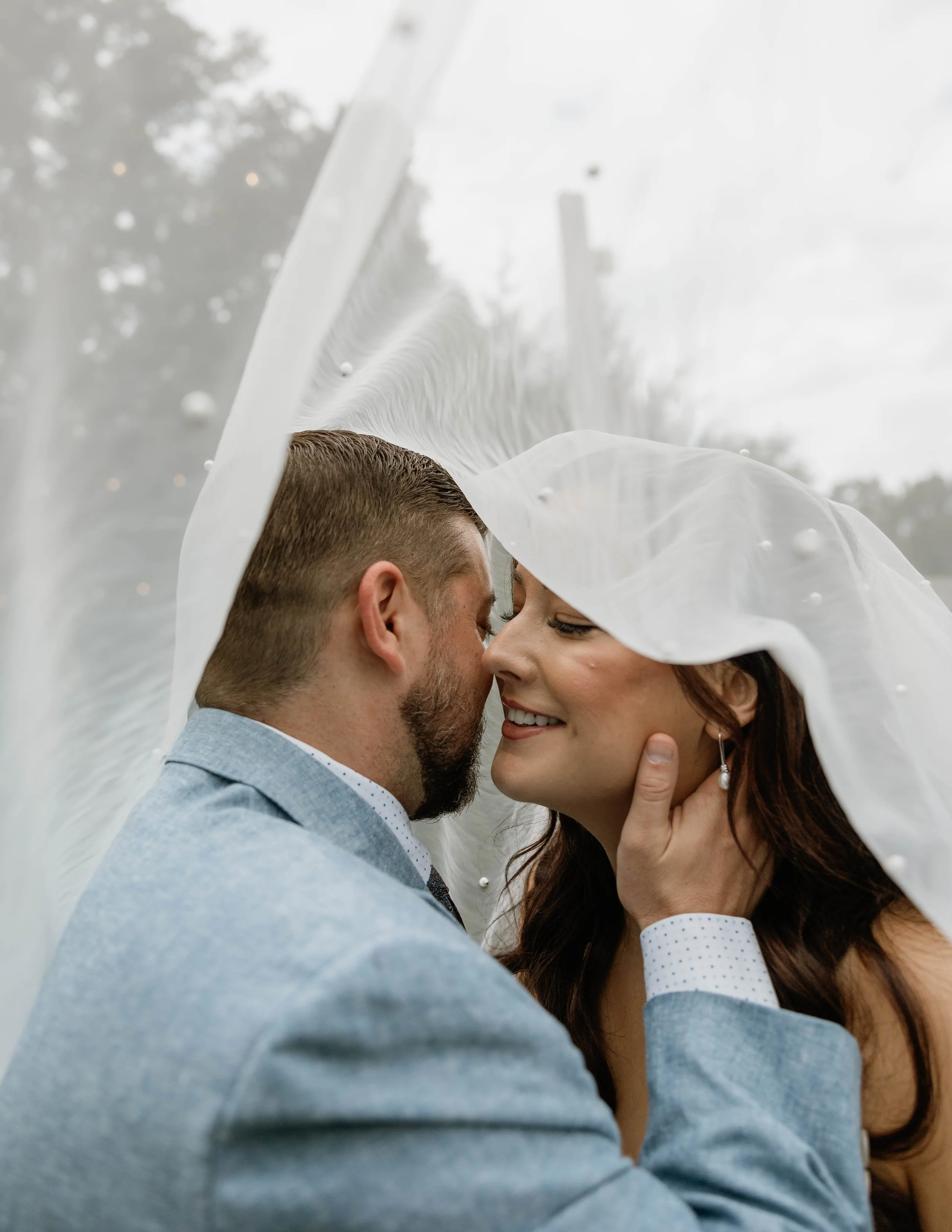 A couple under a wedding veil, sharing an intimate moment outdoors on a cloudy day.