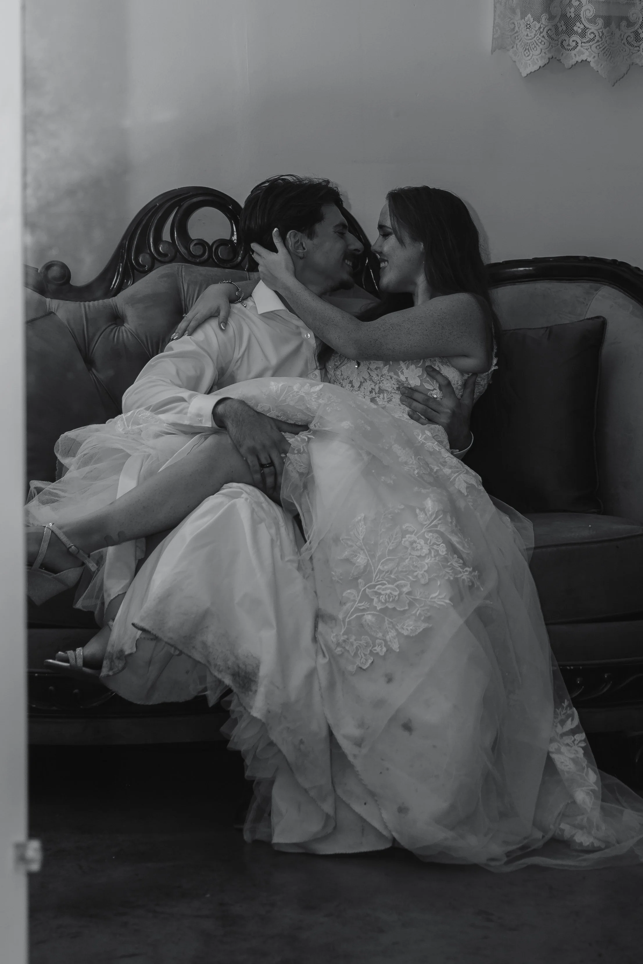 A couple dressed in wedding attire sitting on a vintage couch, with the woman sitting on the man's lap, both smiling and about to kiss.