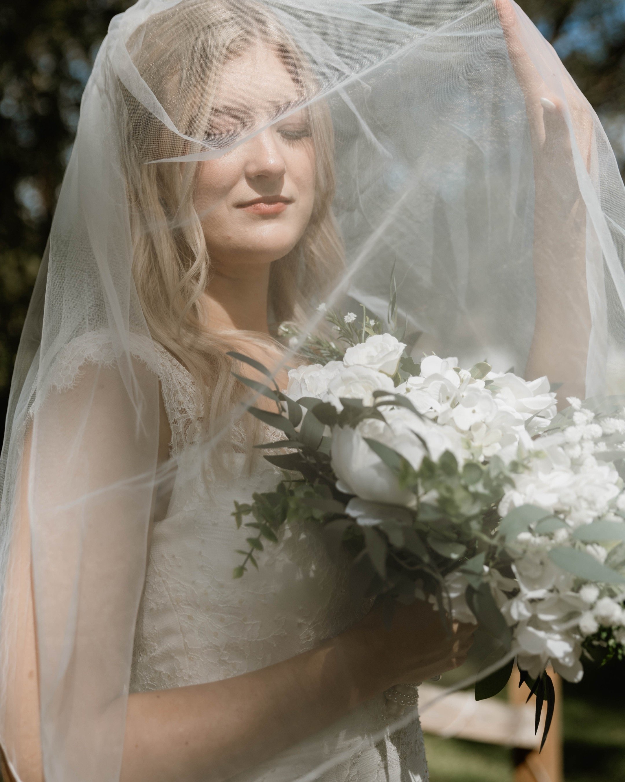A bride with blonde hair and a veil, holding a bouquet of white flowers and green leaves, outdoors with trees in the background.