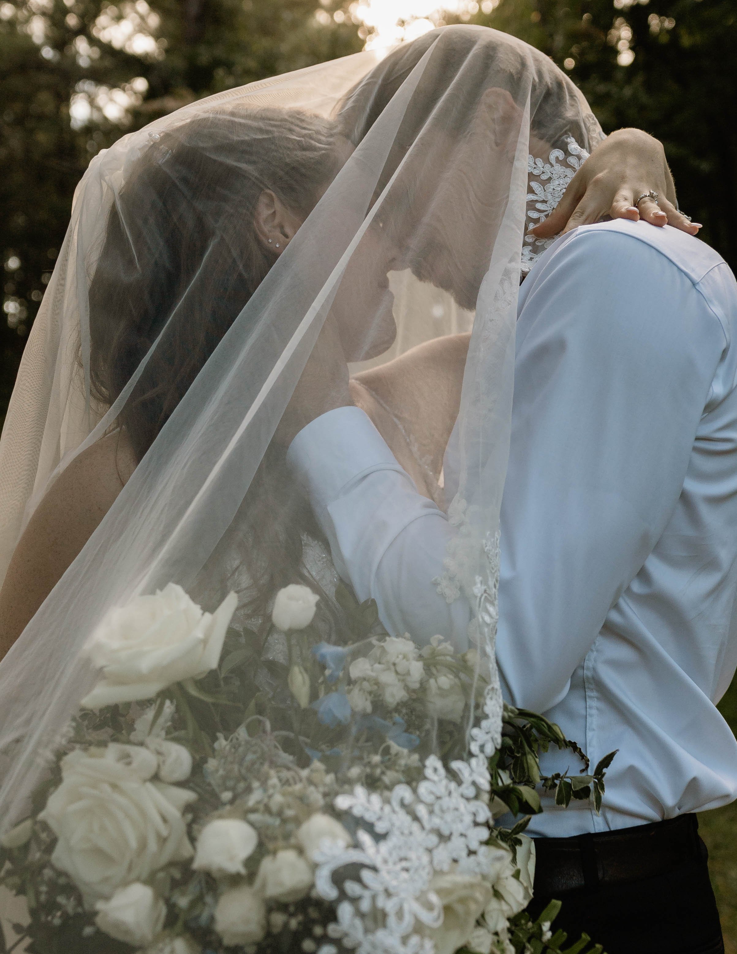 A bride wearing a veil hugs a groom outdoors during sunset, holding a bouquet of white and blue flowers.