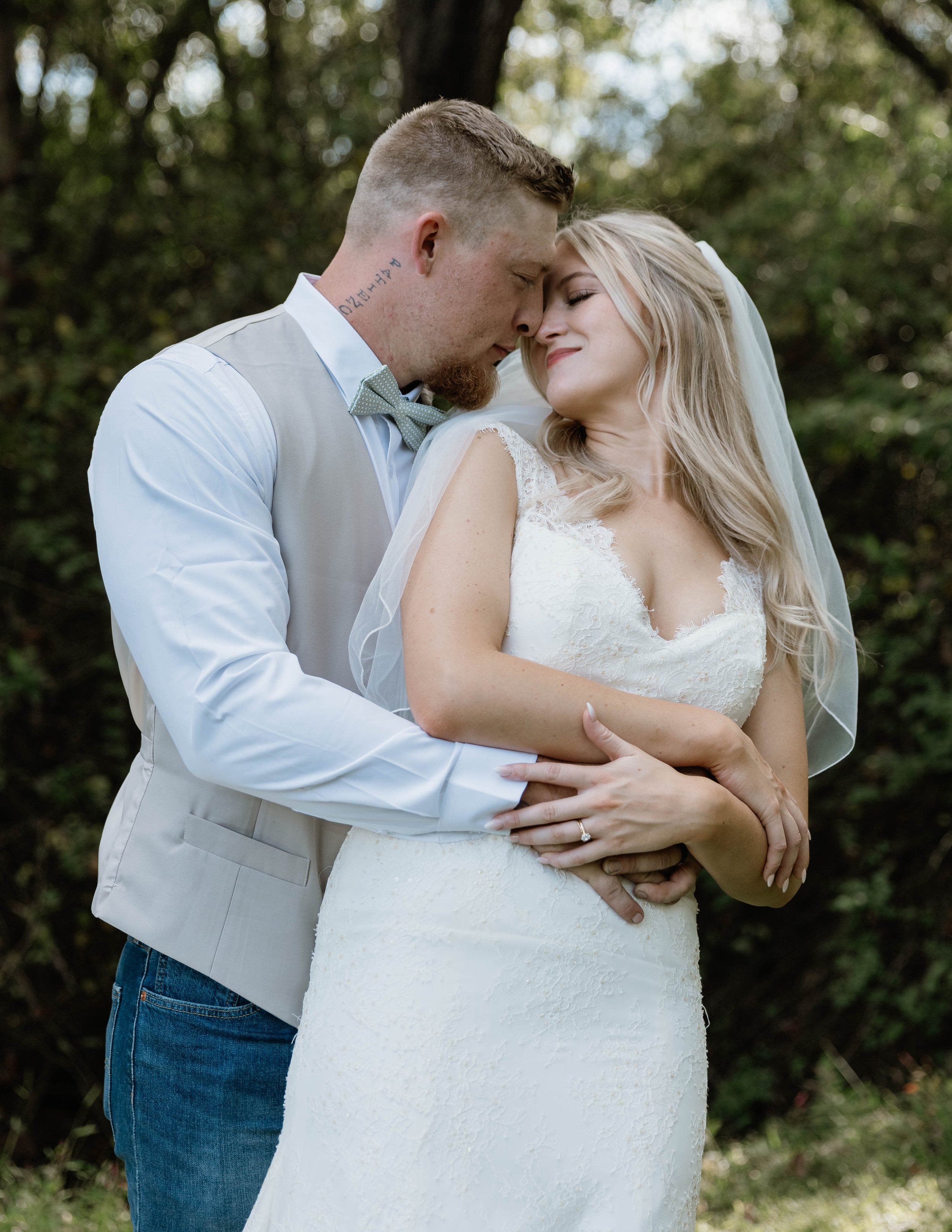 A bride and groom embrace outdoors with trees in the background, eyes closed, touching foreheads, smiling gently, with the groom wearing a light vest and bow tie, and the bride in a lace wedding gown and veil.