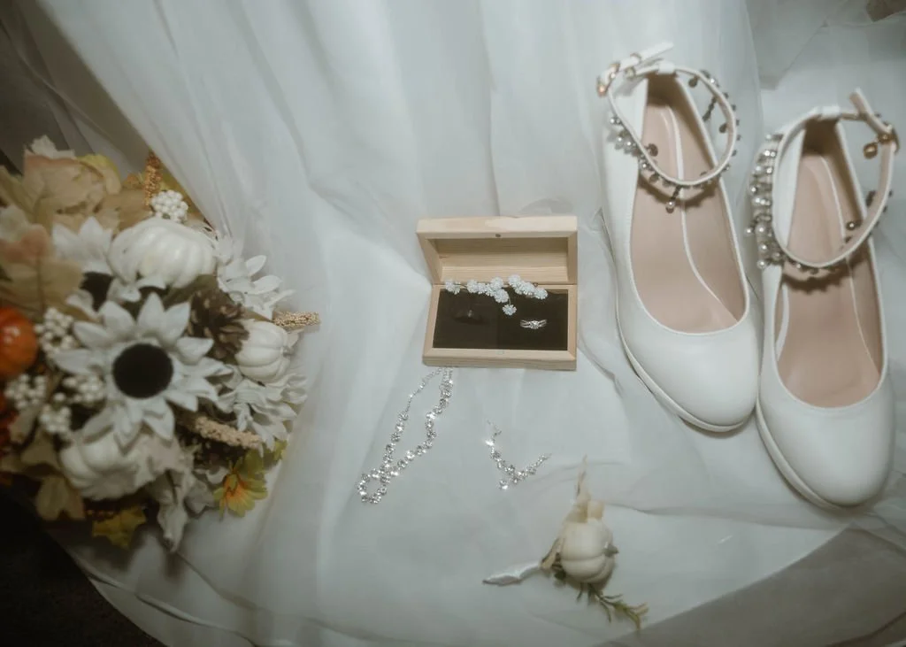 Wedding accessories including white shoes with pearl accents, a pearl necklace, floral bouquet, a wooden jewelry box with rings and earrings, and a white corsage, all arranged on white fabric.