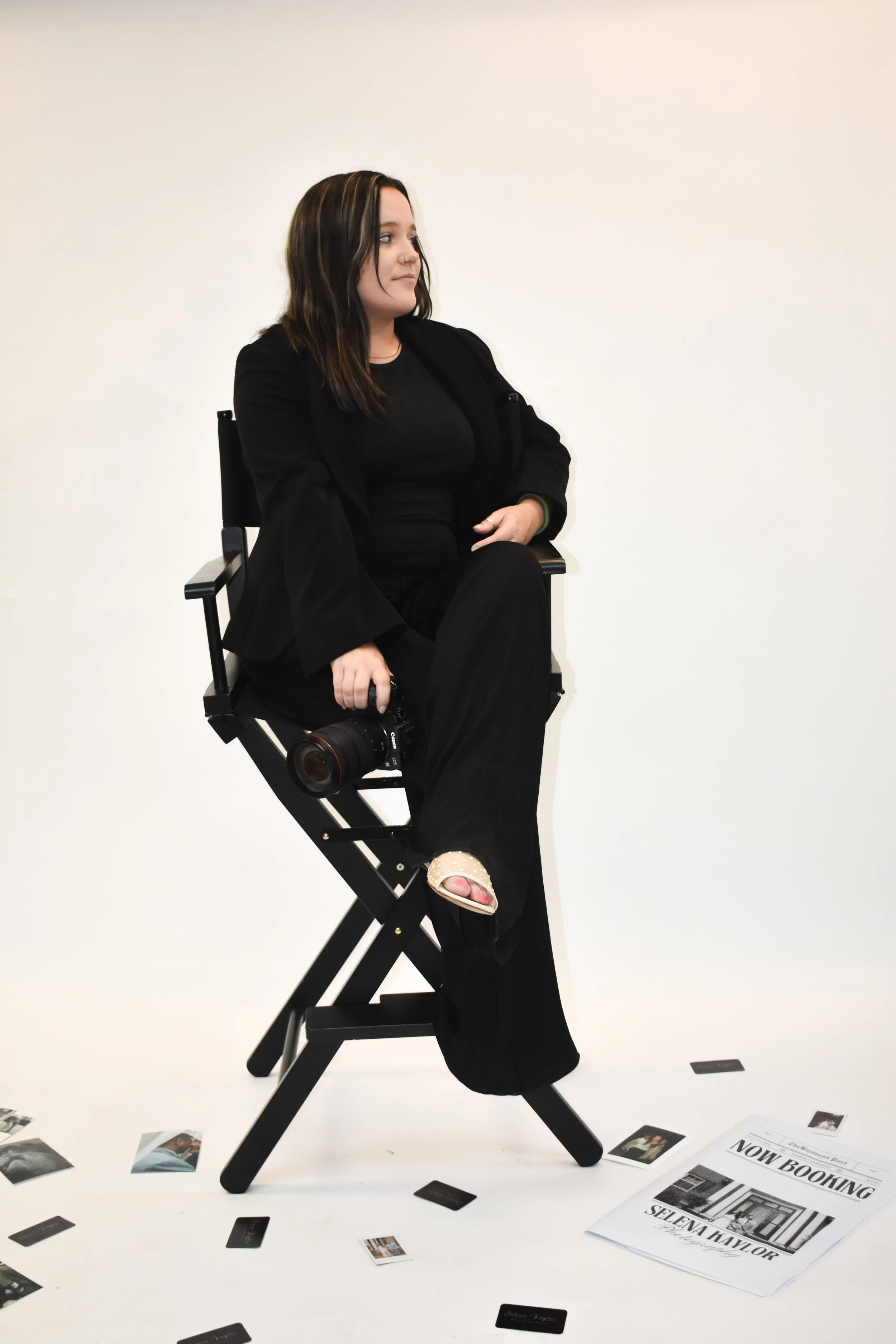 A woman in black clothing sitting on a director's chair, holding a camera, with photos and a flyer on the floor that says 'Now Booking Selena Kaylor'.