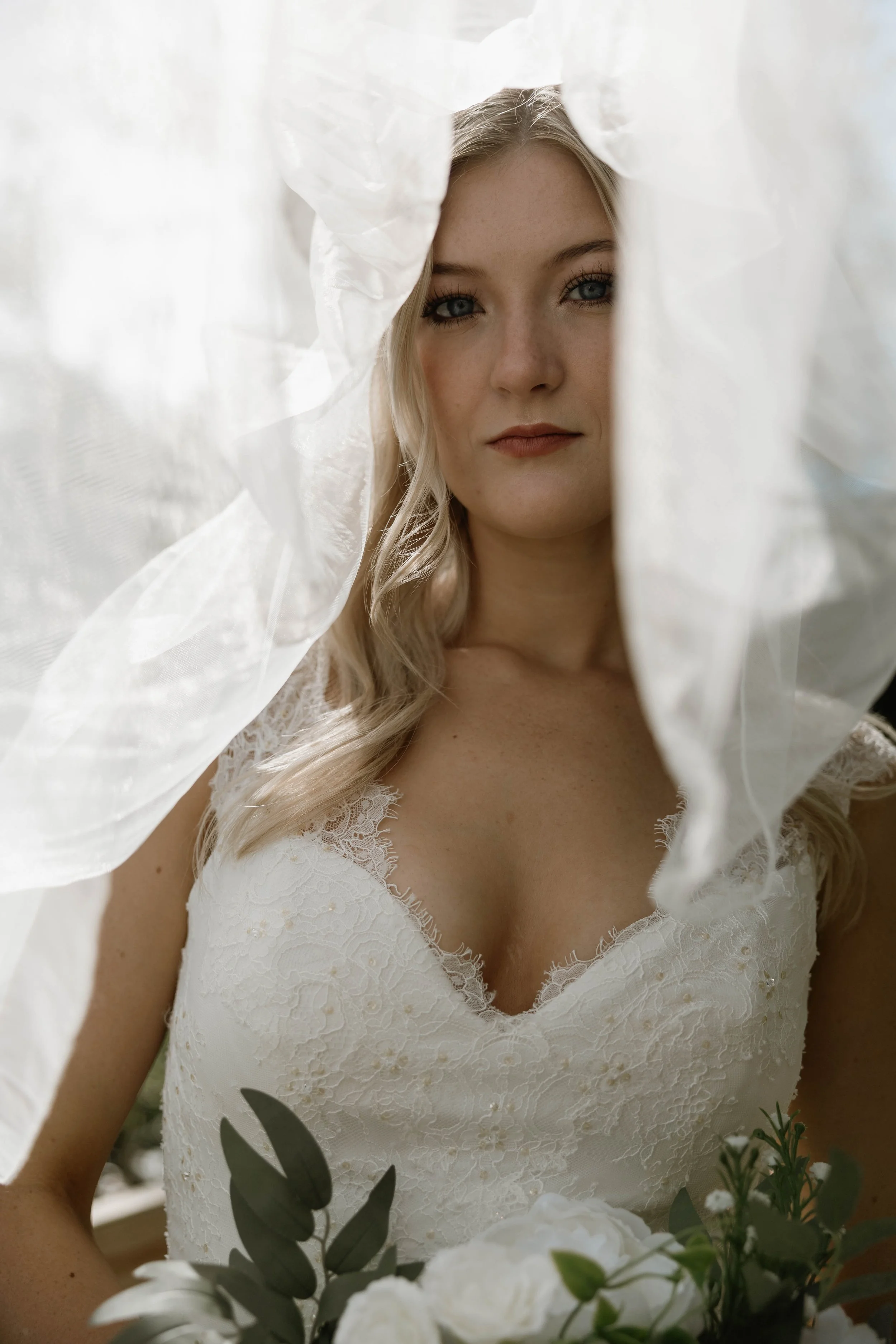 A woman in a white lace wedding dress holding a bouquet of white flowers and greenery, partly obscured by sheer white fabric.