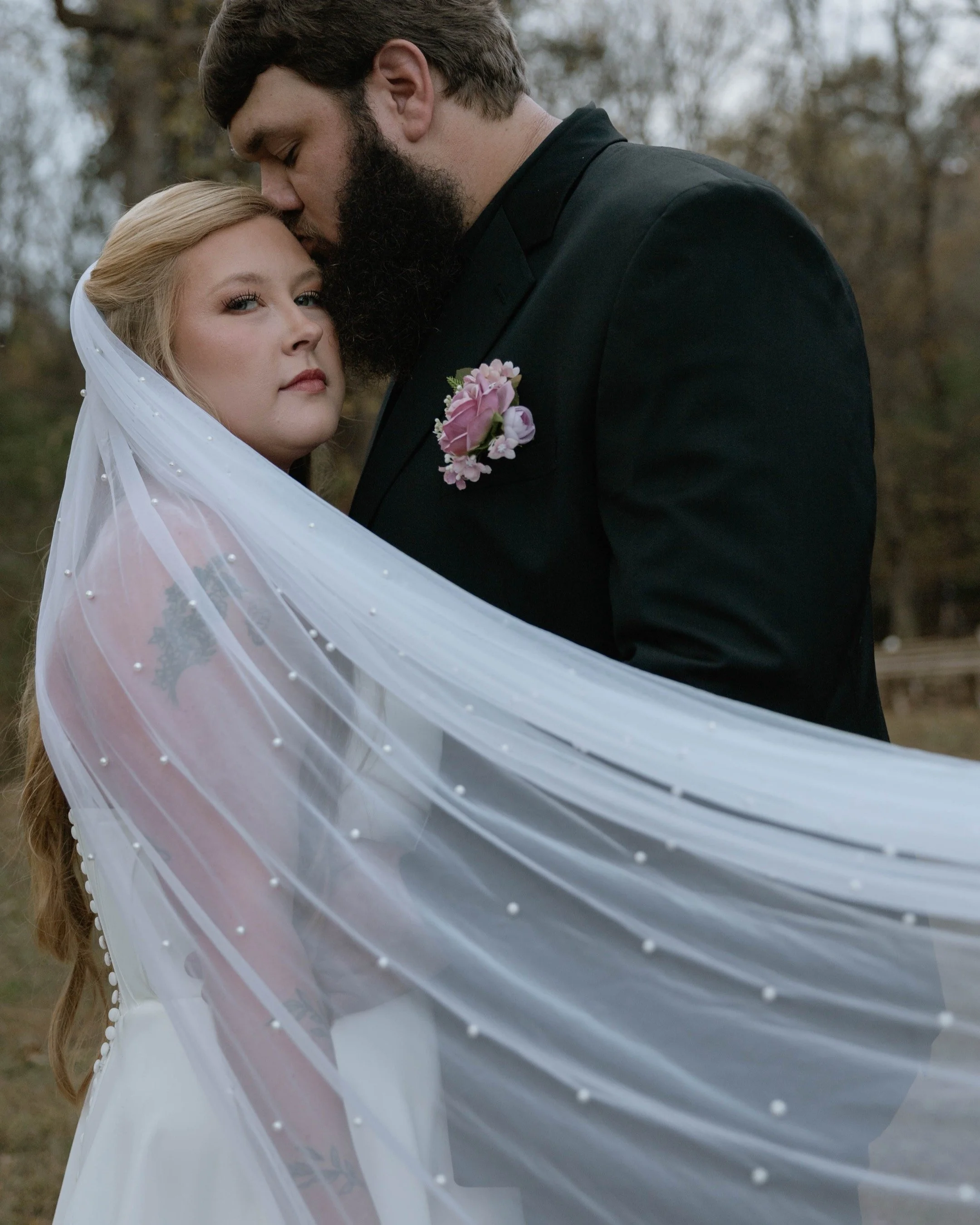 A bride and groom embrace outdoors during a wedding. The bride wears a veil and has blonde hair, and the groom is dressed in a black suit with a pink boutonniere. The bride has tattoos on her arm and a pink flower tattoo on her shoulder.