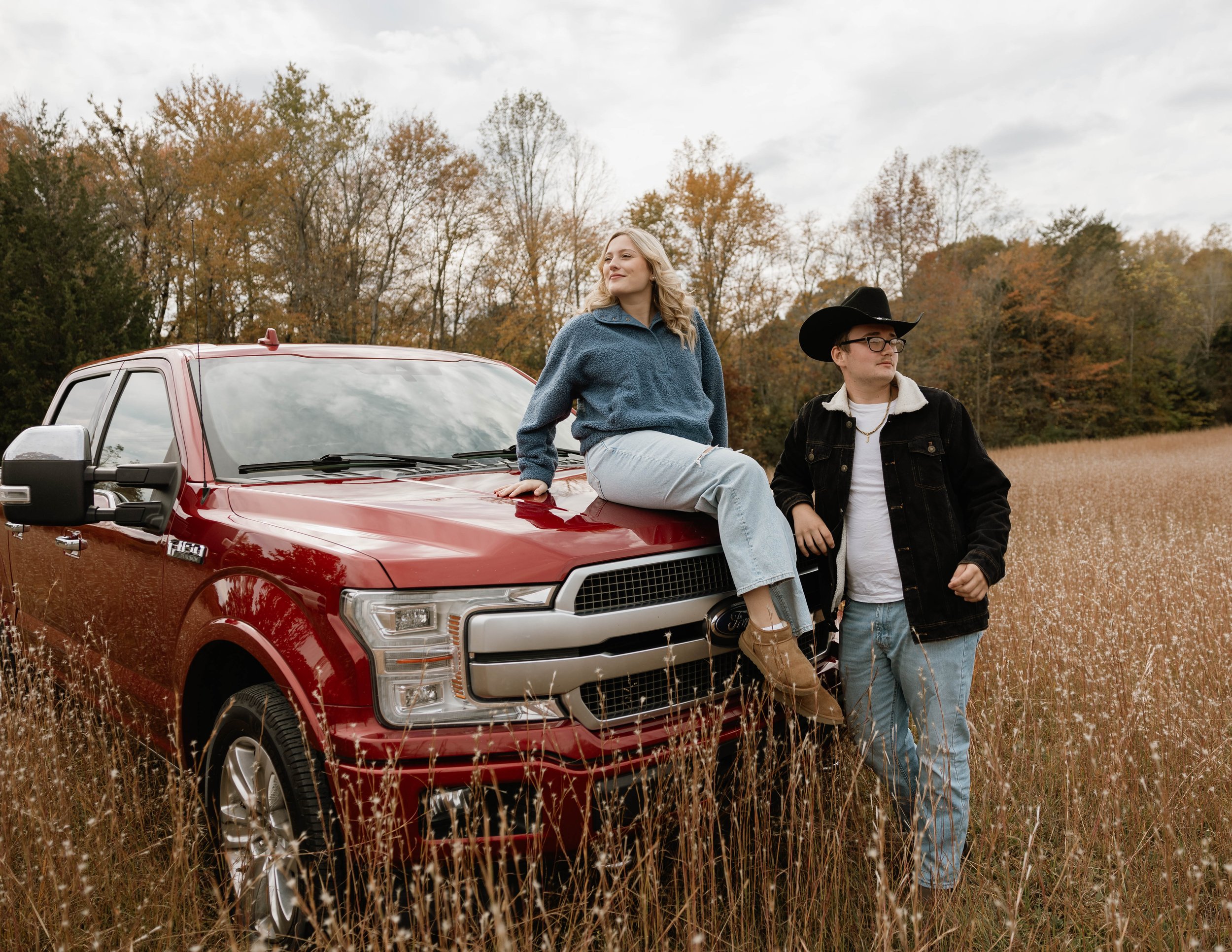 A young woman sitting on the hood of a red truck in a field of tall grass, with a young man standing nearby dressed in a cowboy hat and jacket, with autumn trees in the background.