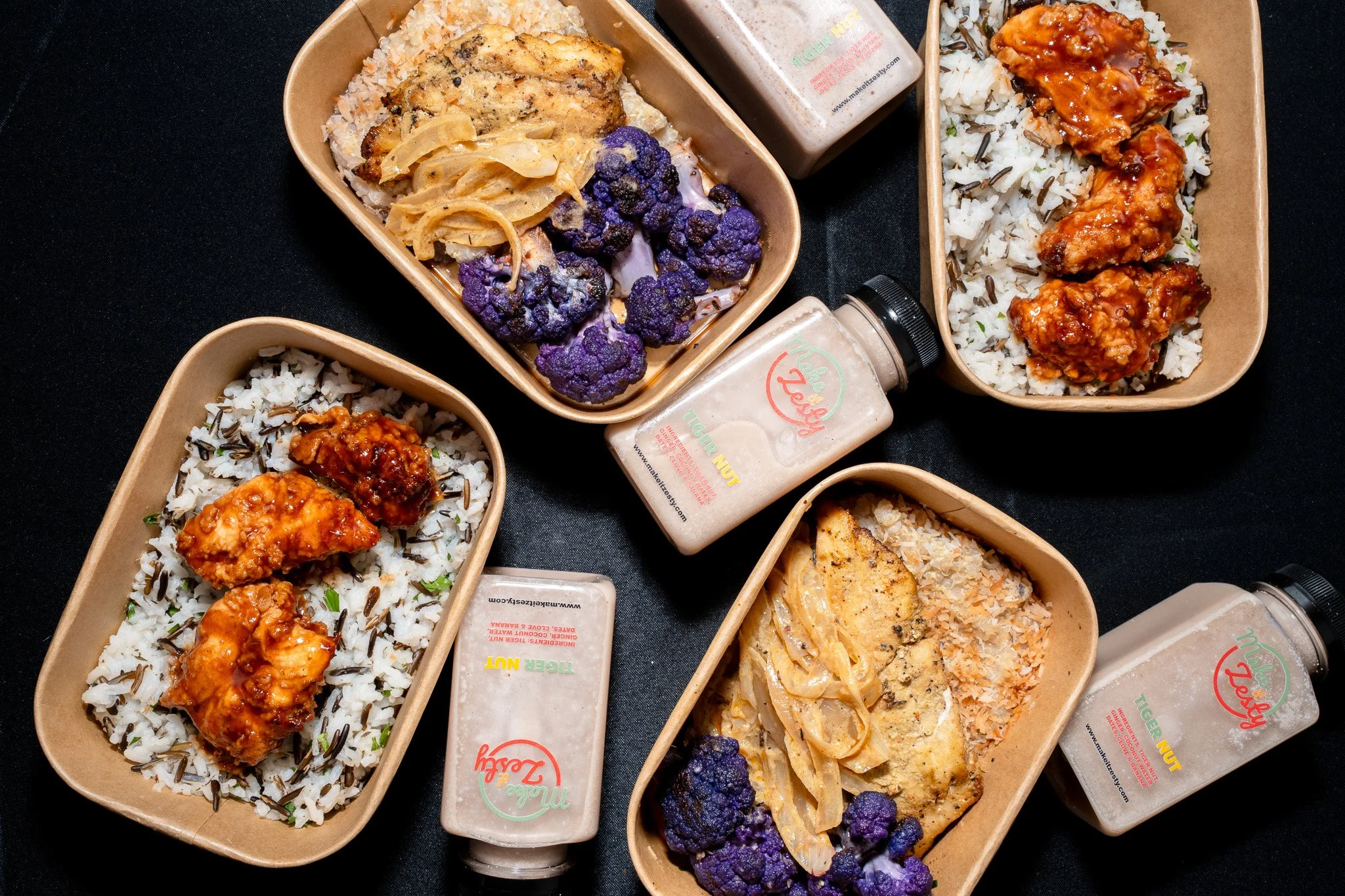 Multiple brown food boxes containing various rice dishes, including rice topped with chicken and sauce, rice with black rice and chicken, and rice with black rice and purple cauliflower, with bottles of seasoning or sauce in the background.
