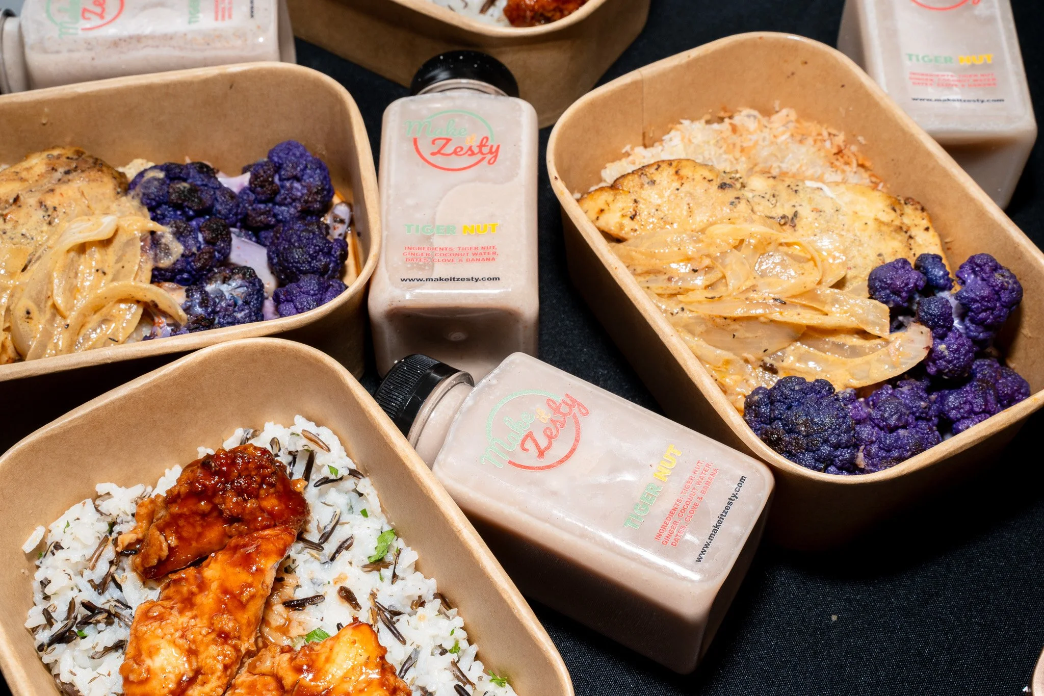 Multiple takeout containers with rice, grilled chicken, purple cauliflower, and various sauces, with bottles of Tiger Nut flavoring in the background.