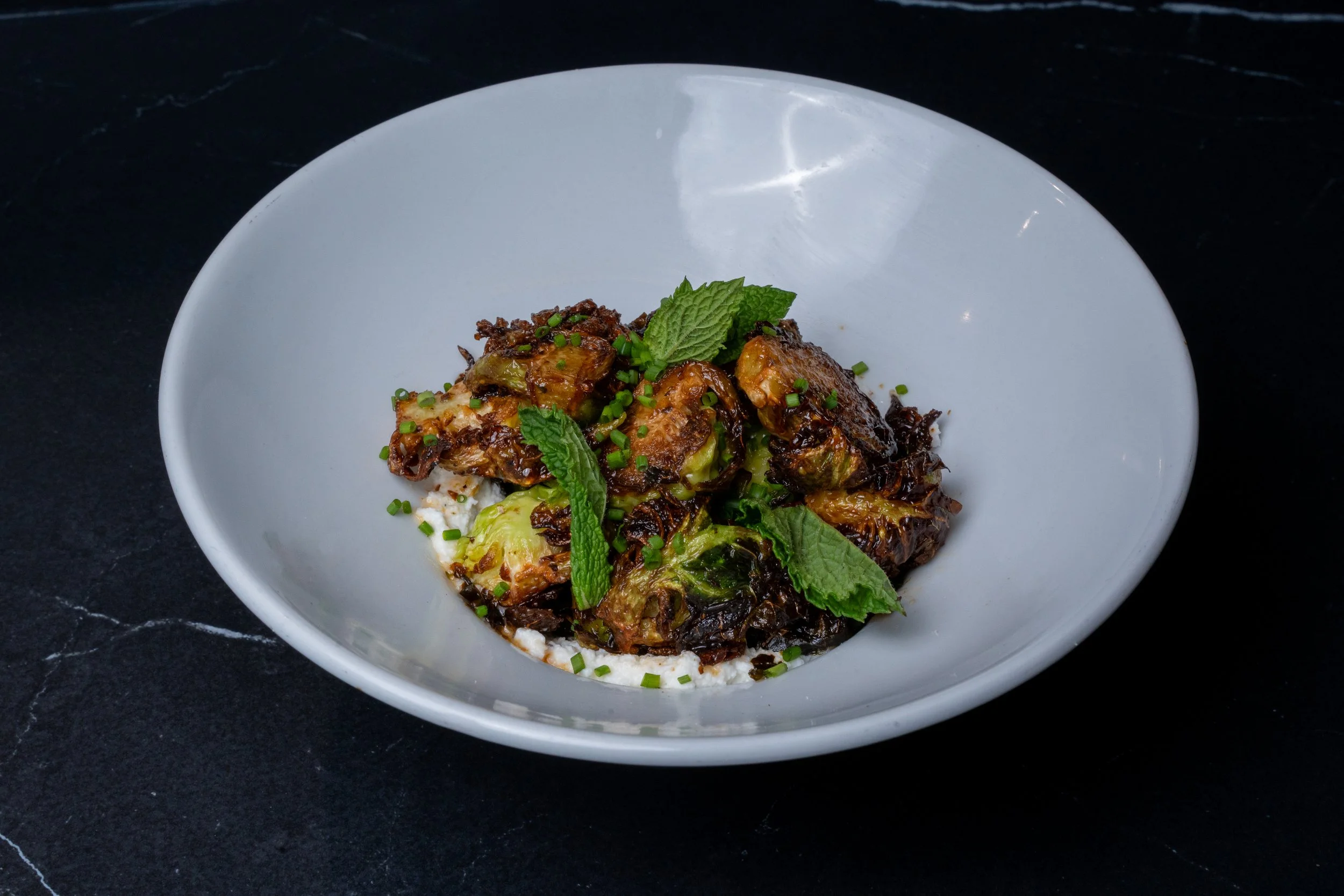 A white bowl containing a dish with cooked vegetables and meat, garnished with fresh mint leaves and chopped green herbs, placed on a black surface.