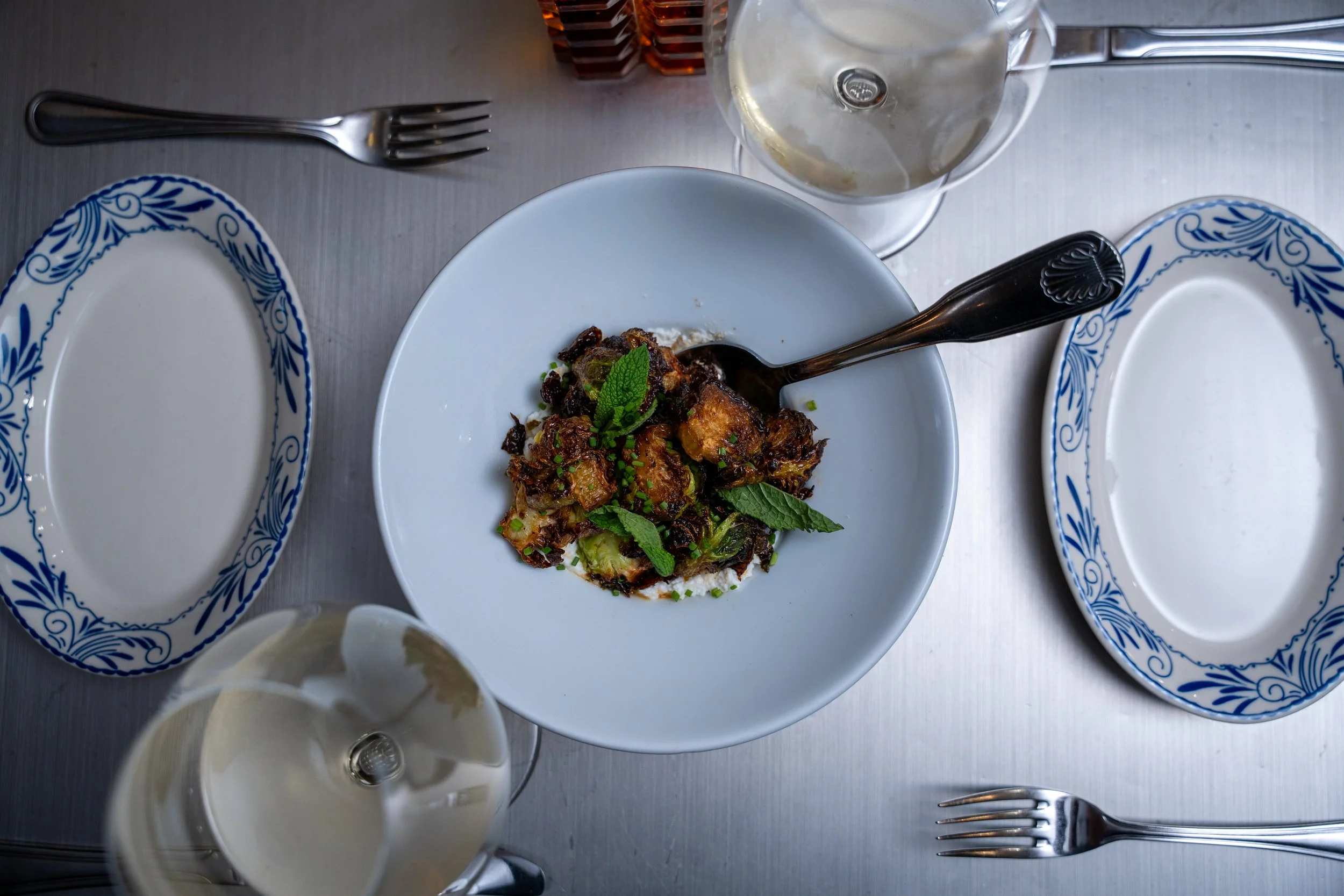 Empty white plate with blue decorative border, a bowl with food garnished with green herbs and a fork, a glass of water, a fork and a knife, and a napkin on a silver table.