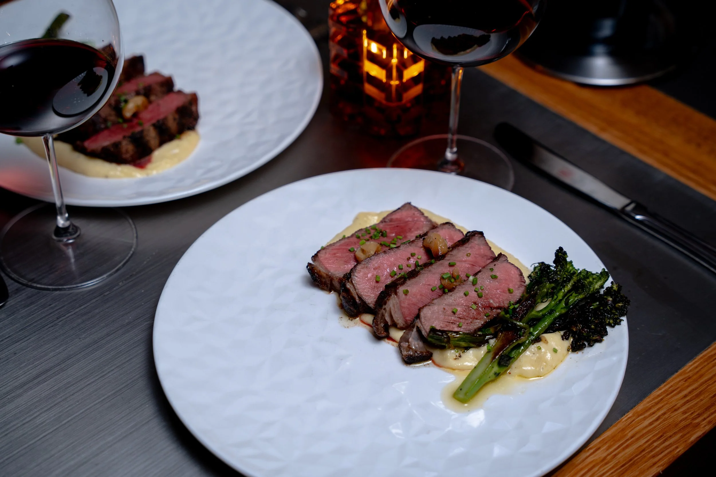A white plate with slices of medium-rare steak topped with chopped chives, served with grilled broccoli and a creamy white sauce, alongside two glasses of red wine, on a dark table with a candle in the background.