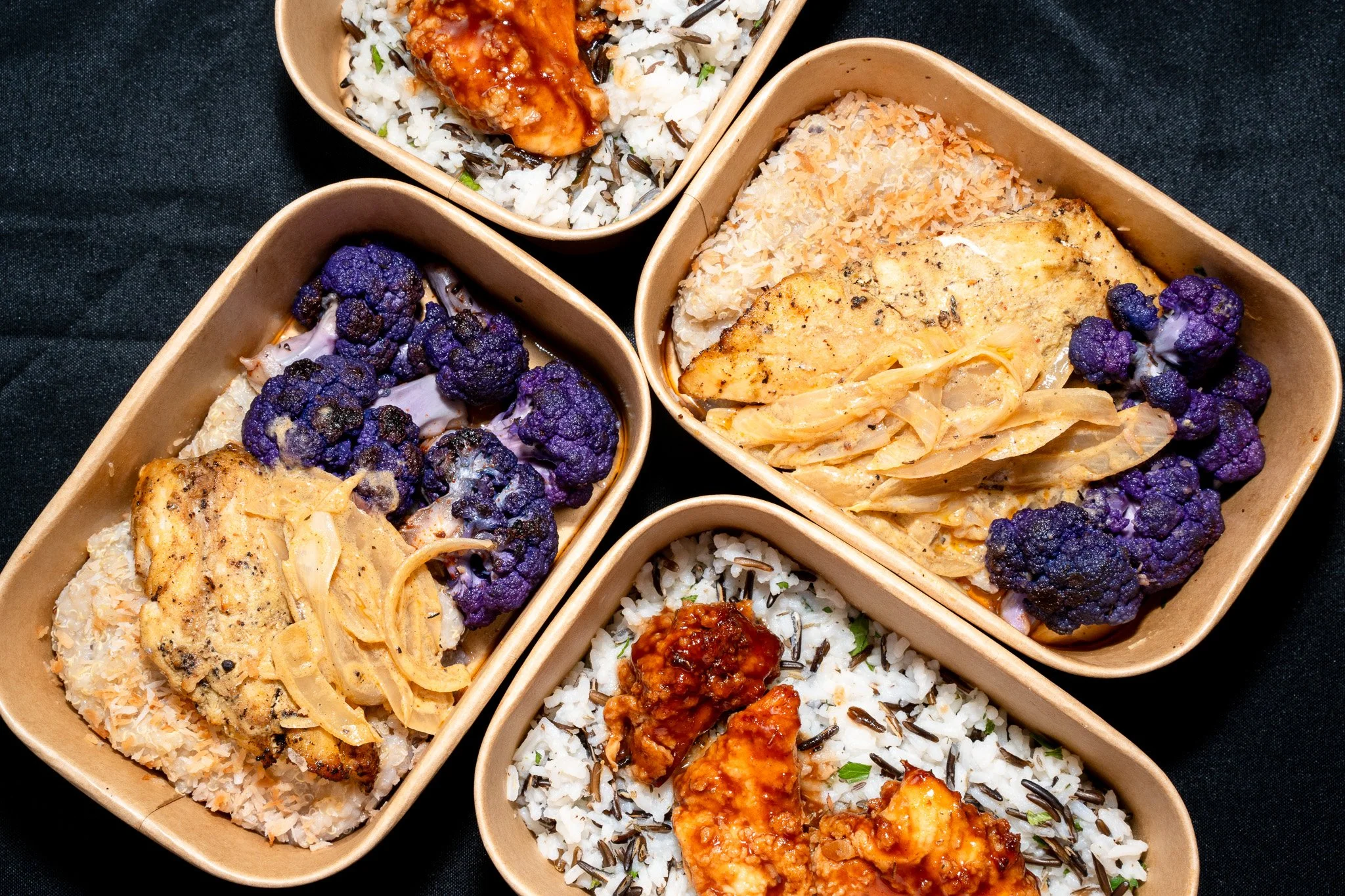 Bento boxes filled with rice, cauliflower, chicken, and vegetables on a black surface.