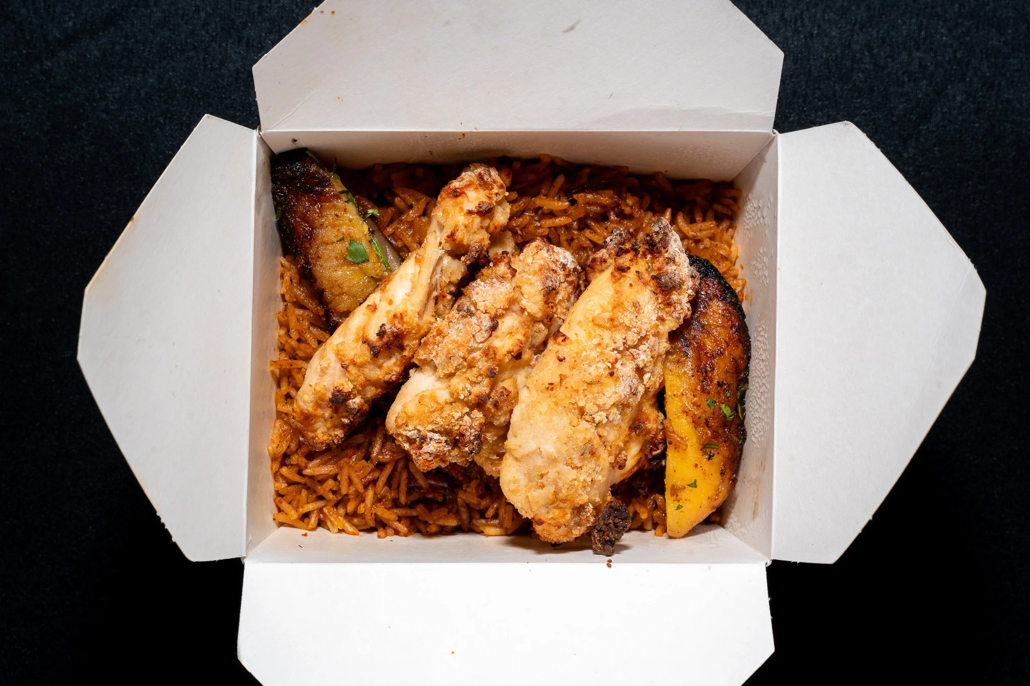 Close-up of a takeout box containing grilled chicken tenders, fried plantains, and seasoned rice, placed on a black surface.