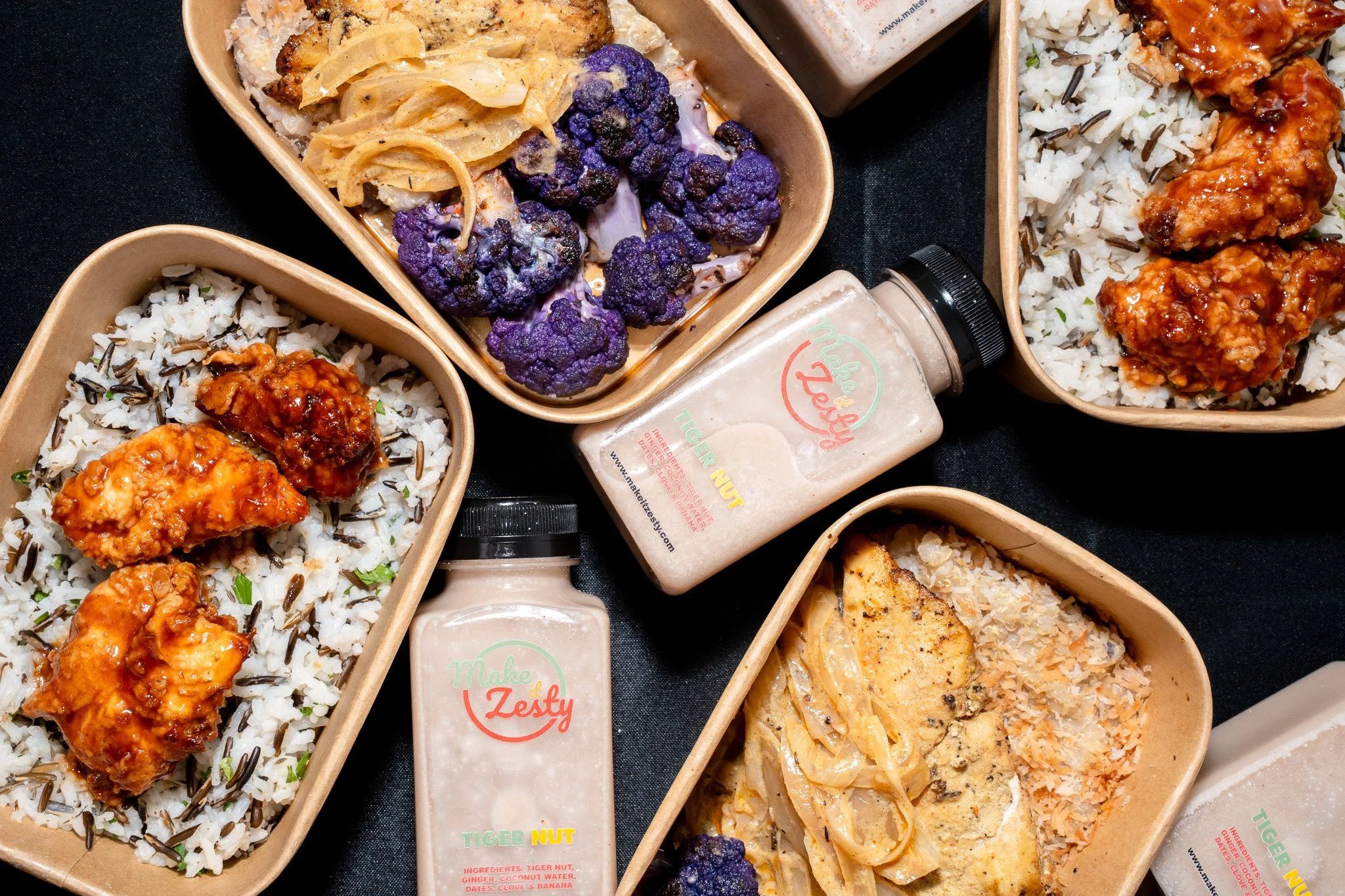 Takeout containers with rice, grilled chicken, purple cauliflower, and pasta dishes, accompanied by bottles of sauces on a black surface.