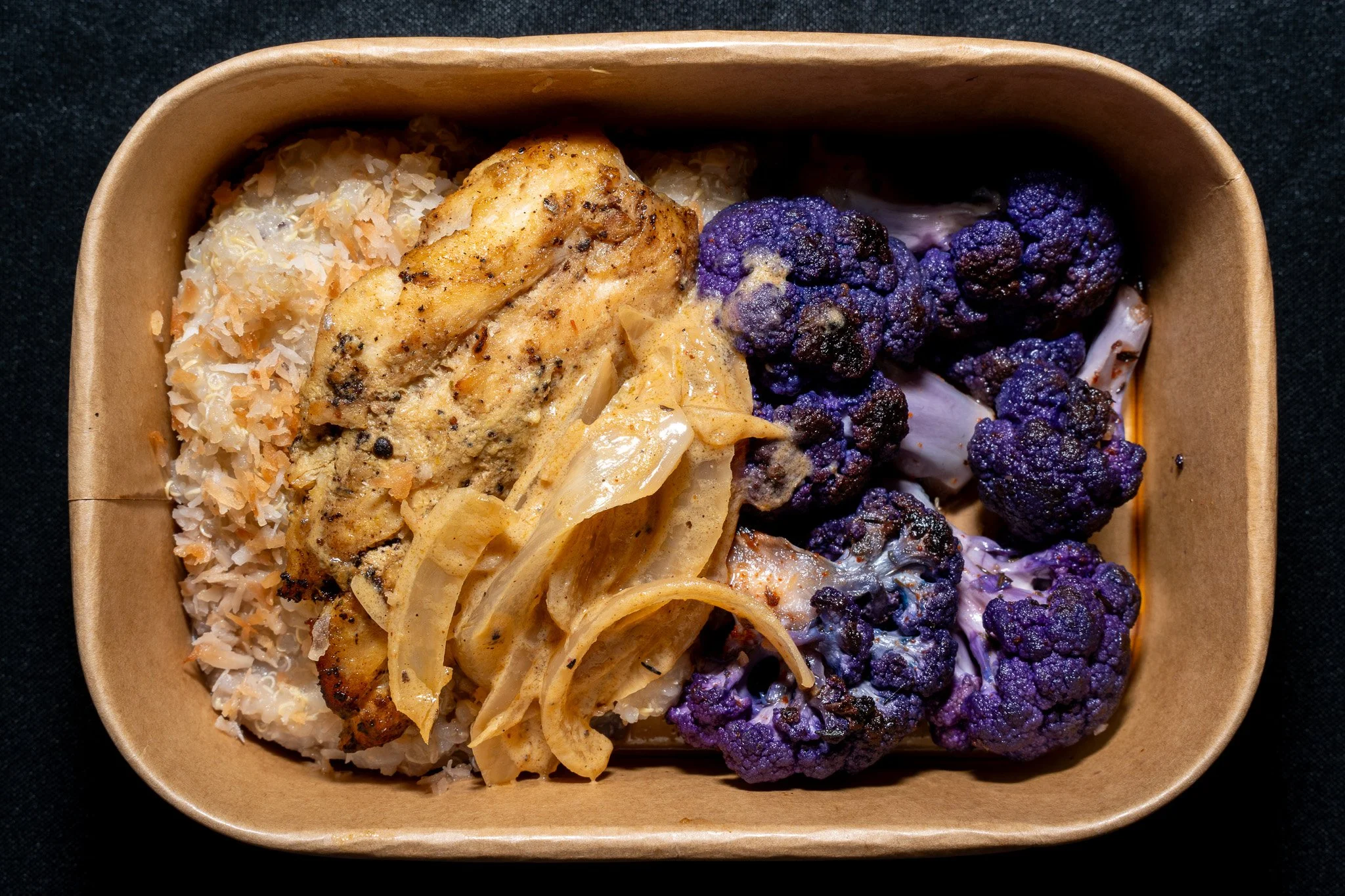 A brown paper food tray with cooked cauliflower florets, grilled chicken breast, cooked onions, and sauced pasta, served on a black surface.