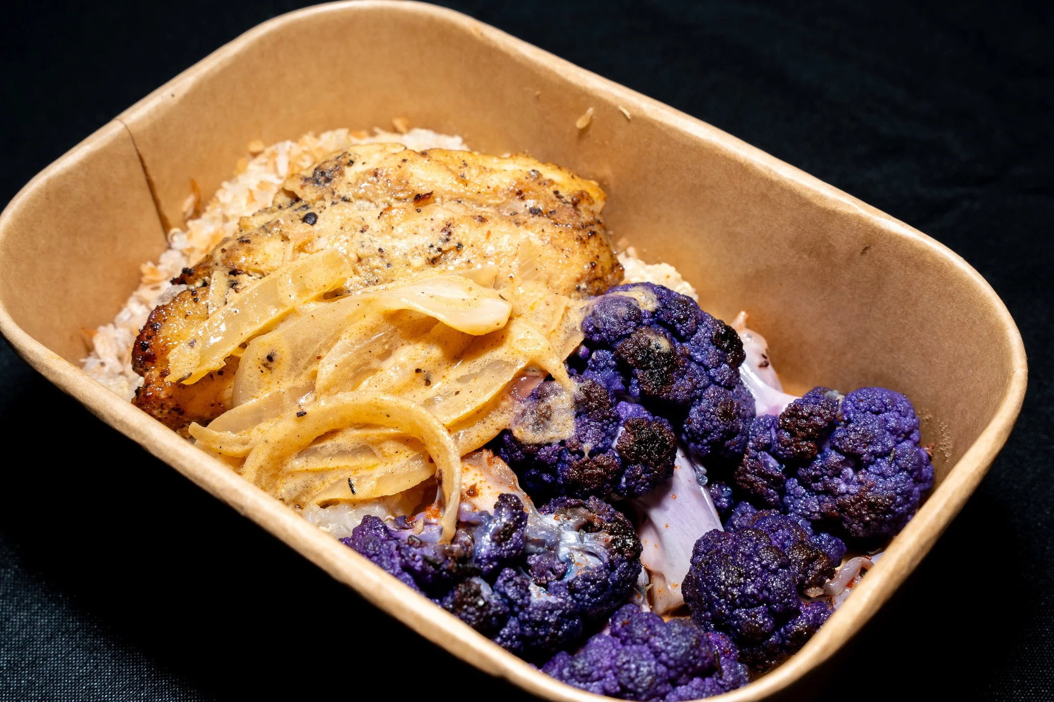 A brown takeout container filled with roasted chicken, pasta with sauce, and purple cauliflower florets.