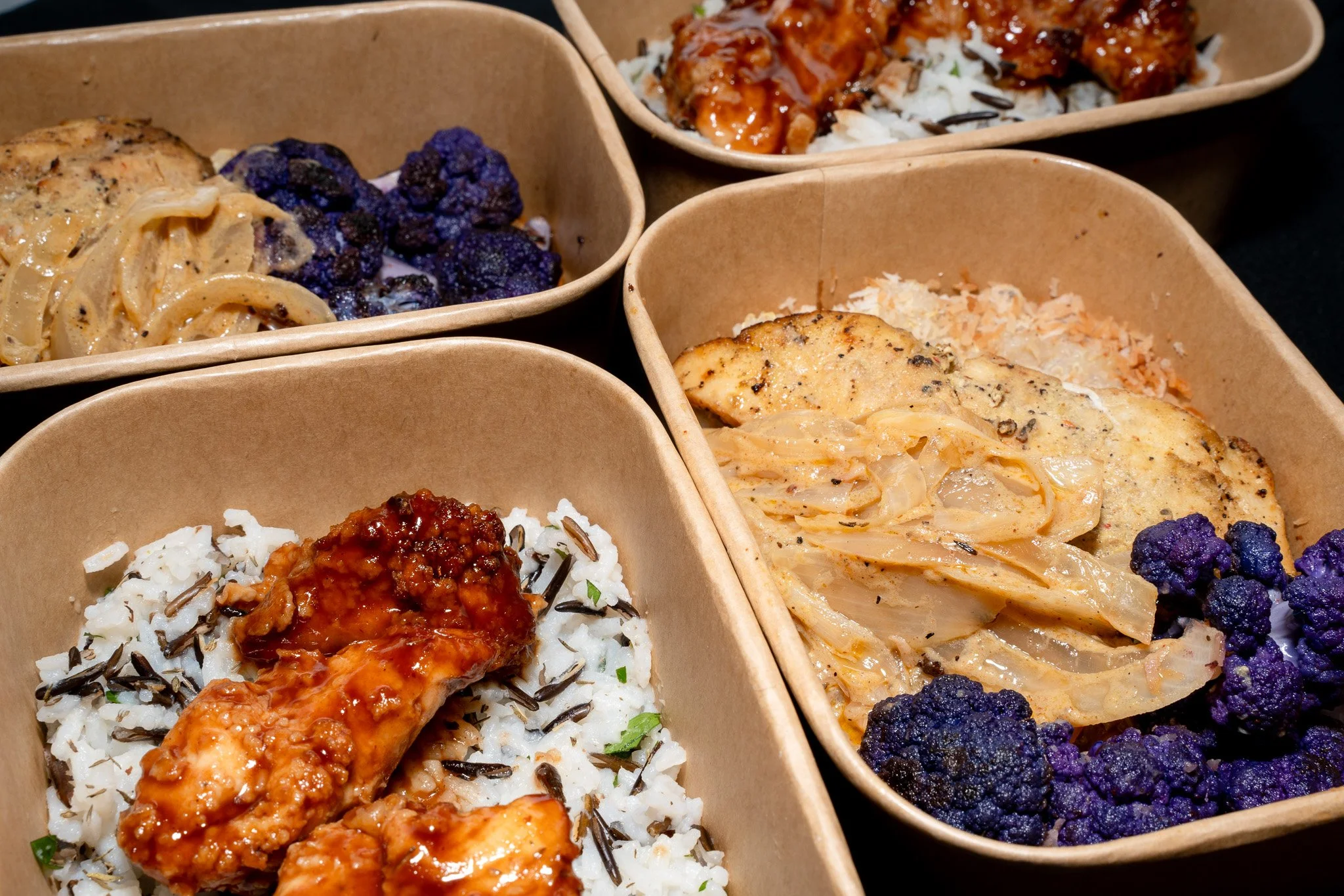 Three disposable food containers filled with assorted rice and chicken dishes, including black rice, topped with sauces and vegetables.