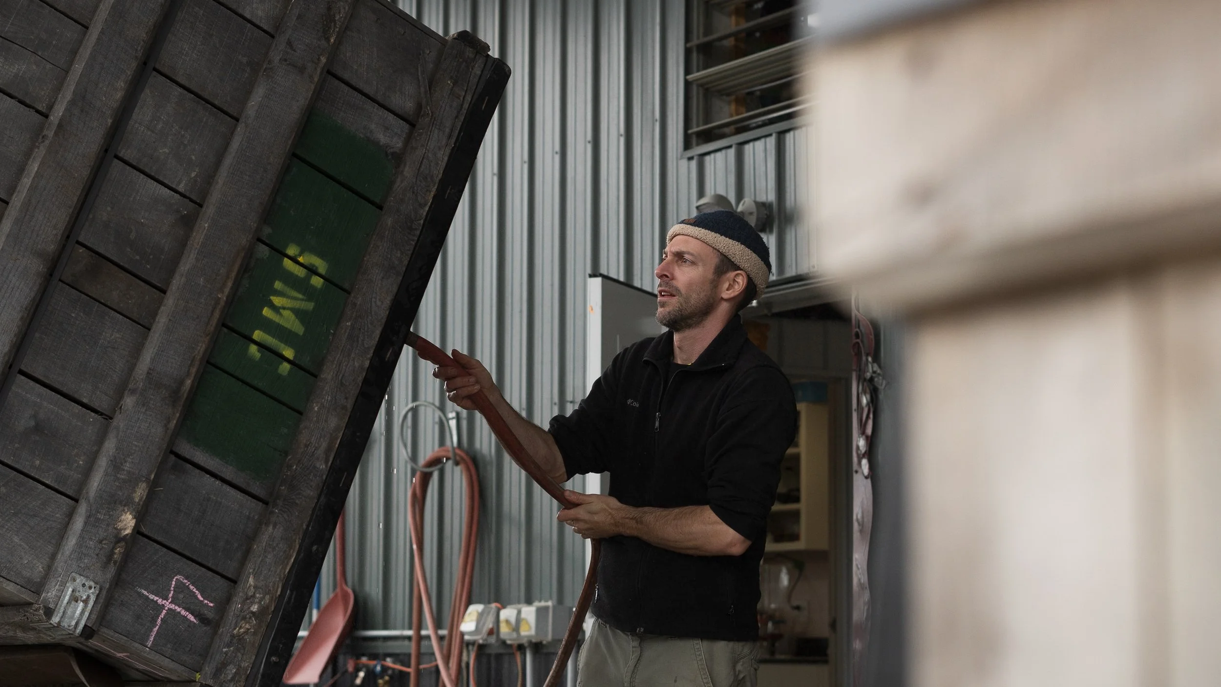 A man with a beard and wearing a beanie hat is working inside a workshop or storage area, examining or repairing an old, weathered wooden crate or fence panel.