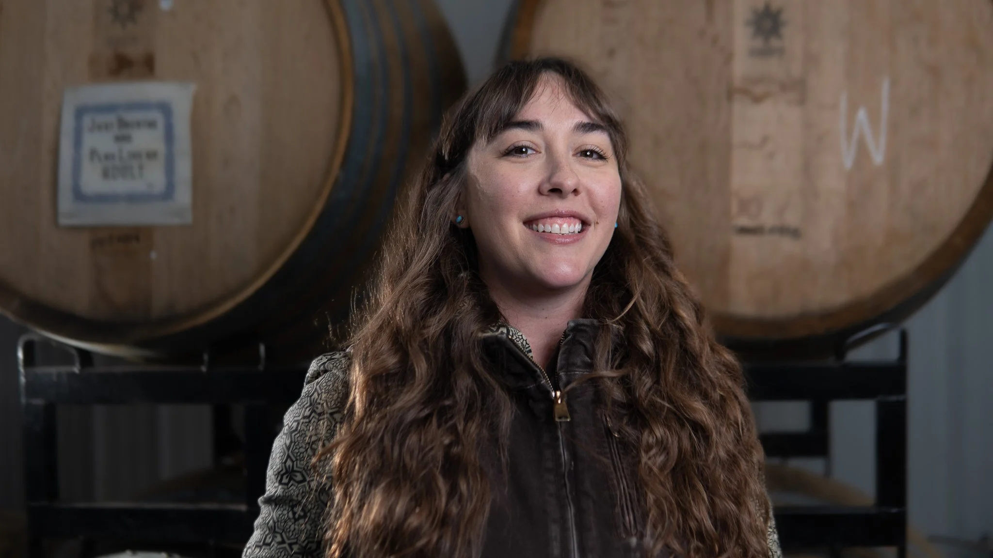 Woman with long curly hair smiling in a brewery or winery with large wooden barrels in the background.
