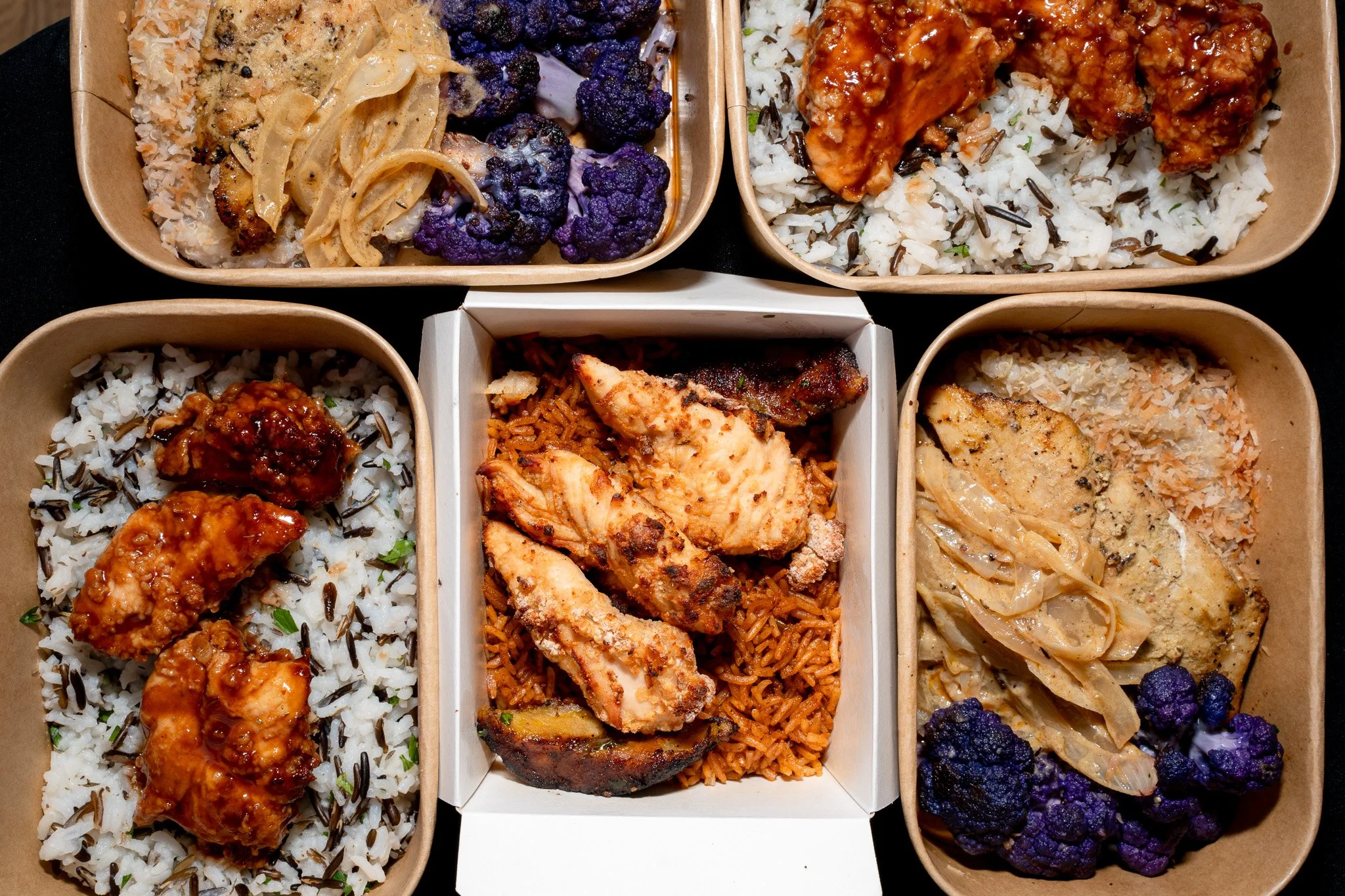 Multiple compartments with various types of rice dishes including white rice with black rice, spicy fried chicken, and roasted chicken, accompanied by purple cauliflower florets and creamy pasta.