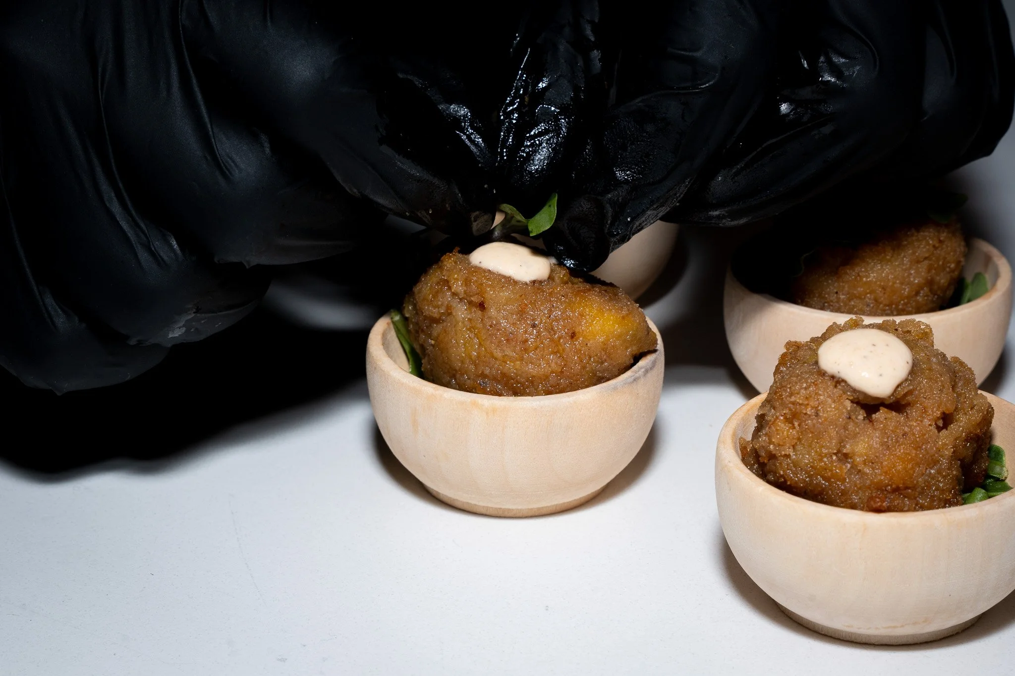 A person wearing black gloves placing a dollop of sauce on a round, breaded appetizer served in small wooden cups, with other appetizers in the background.