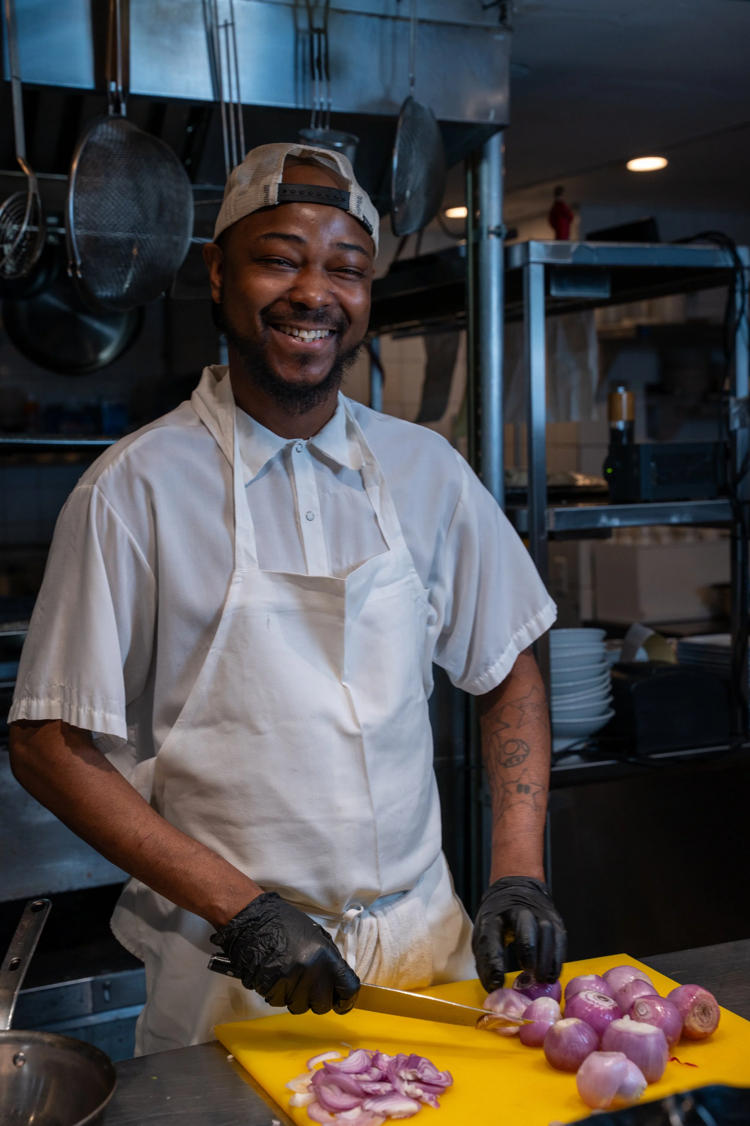 A man in a chef's outfit with a backwards cap and black gloves is slicing purple onions on a yellow cutting board in a commercial kitchen. There are metal kitchen utensils hanging in the background.
