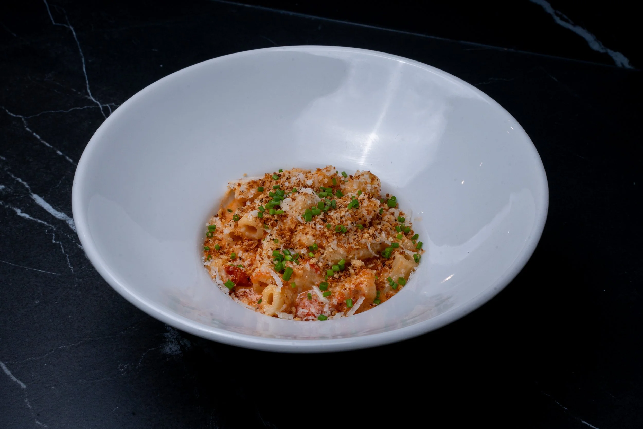A white bowl of cheesy pasta topped with chopped green herbs on a black marble surface.