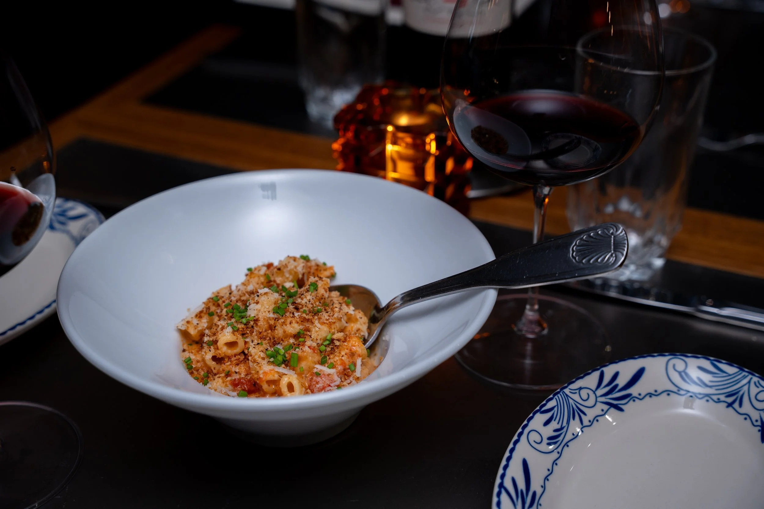 A white bowl of baked pasta with a breadcrumb topping and green chives, placed on a dark table next to a glass of red wine and various plates.