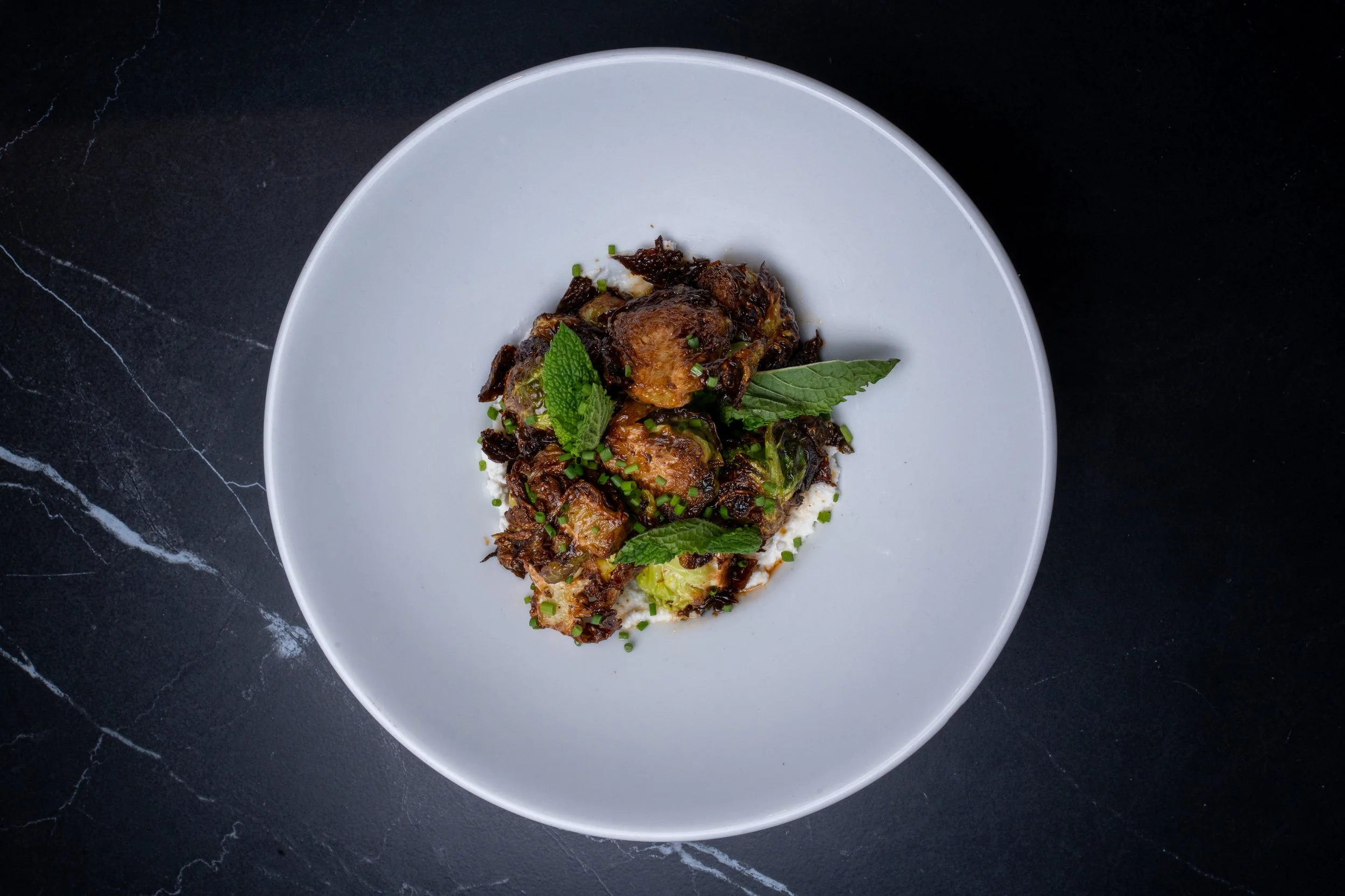 A white plate with roasted chicken and vegetables garnished with fresh mint and chives on a black marble surface.