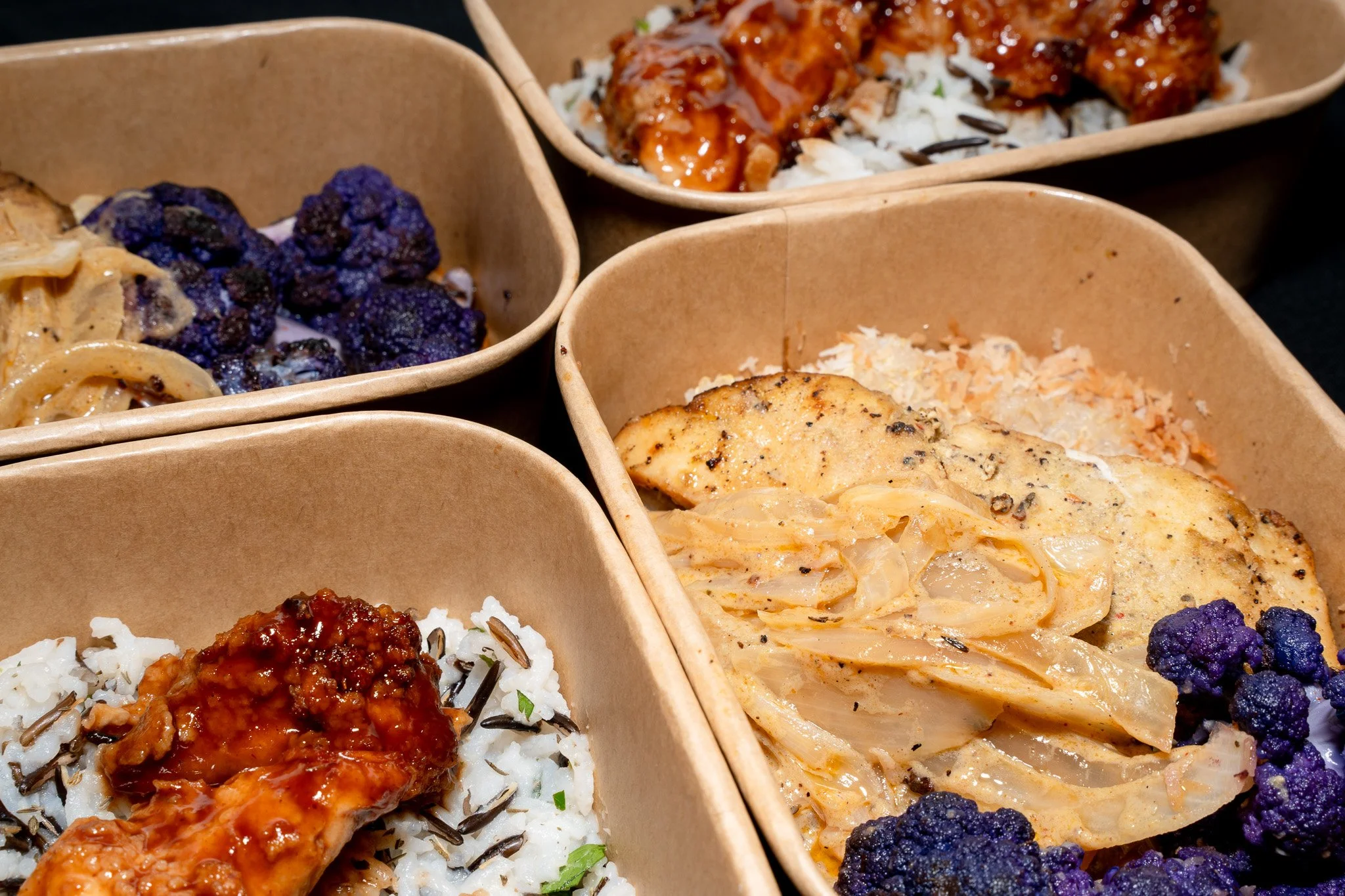 Brown takeout containers filled with rice, chicken, and vegetables, including black and purple cauliflower.