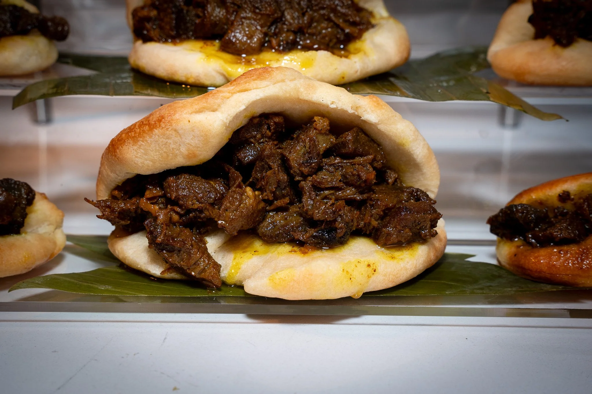 Close-up of a small sandwich bun filled with shredded beef or brisket, on a green leaf with other similar sandwiches in the background.